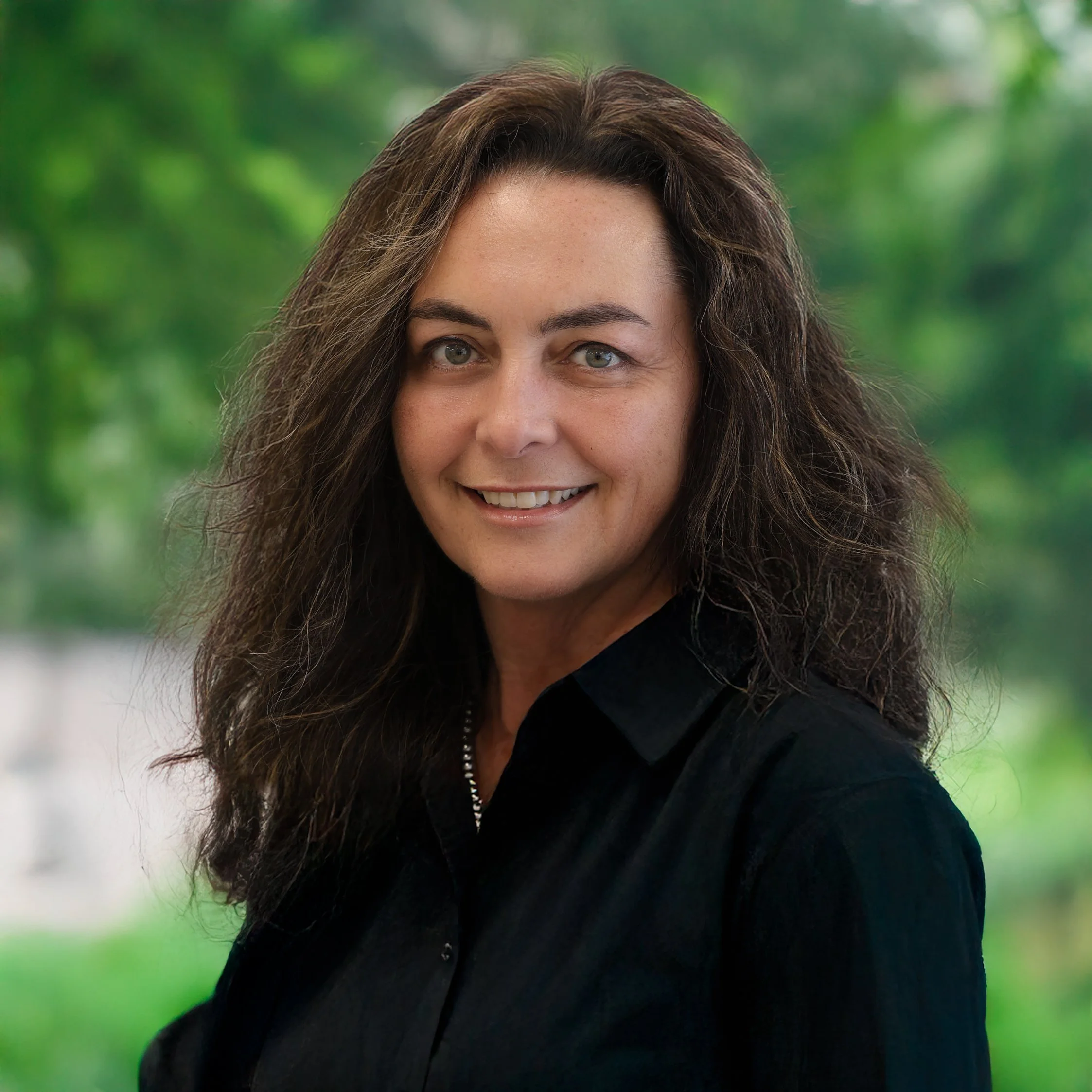 A woman with long curly brown hair wearing a black top, smiling outdoors with a blurred green background.