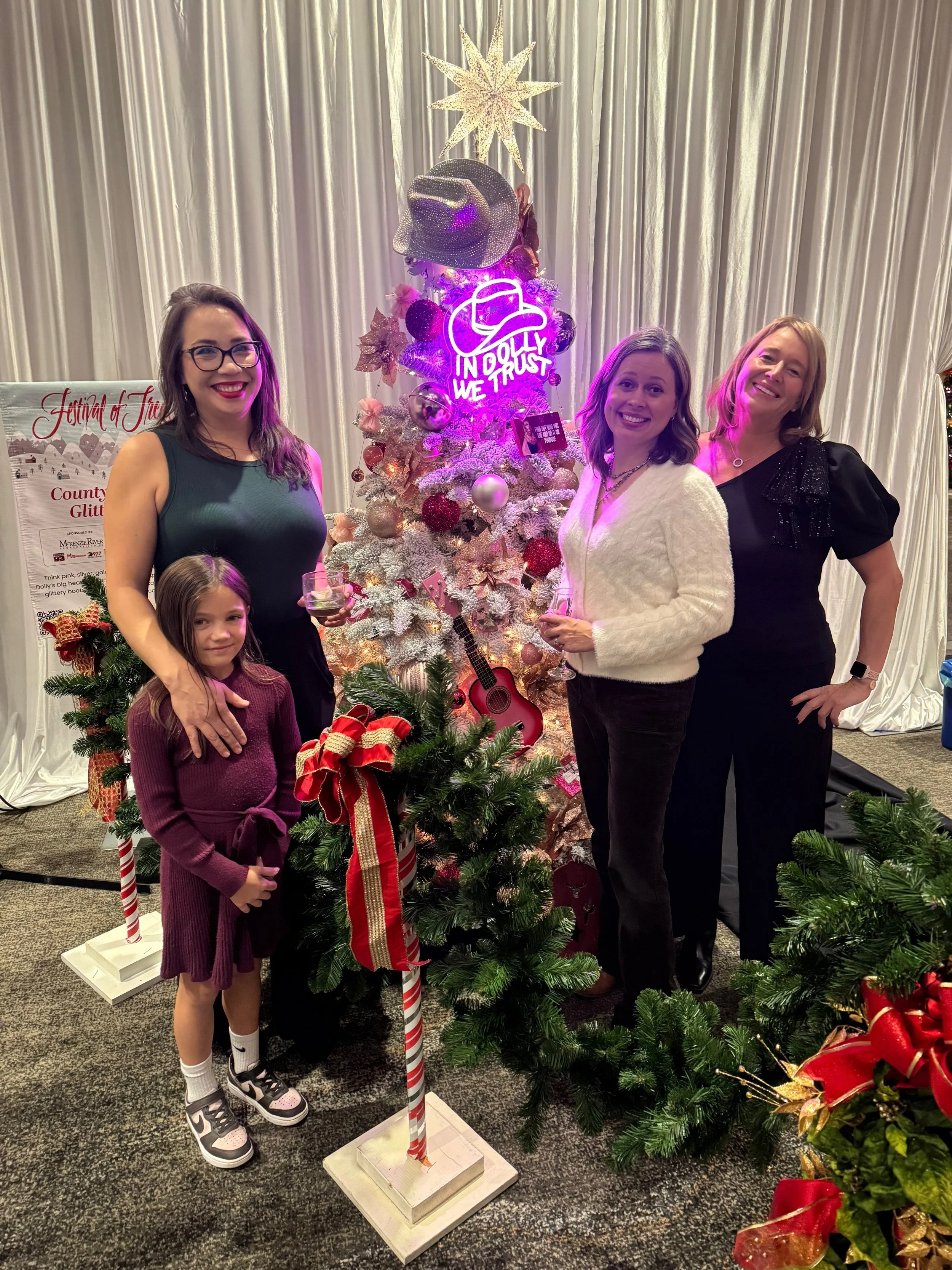 Four women and a girl posing around decorated Christmas trees, with a backdrop of white curtains. One woman holds a wine glass. The Christmas trees have ornaments, ribbons, and neon signs that say 'In Dolly We Trust'. There are star and hat decorations at the top of the tree, and a pink guitar leaning against it.