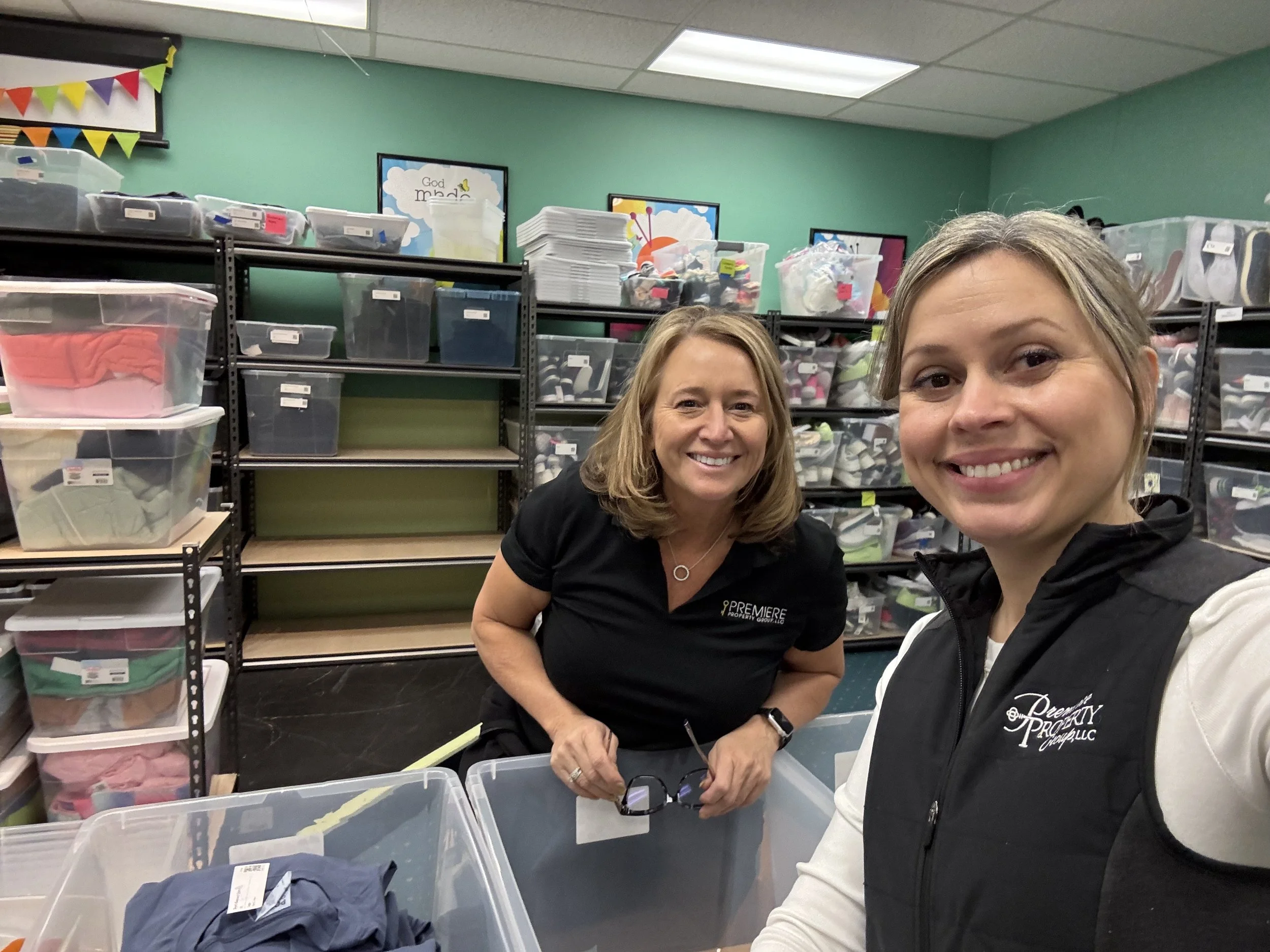 Two women smiling in a storage room with shelves filled with plastic bins of folded clothes in the background.
