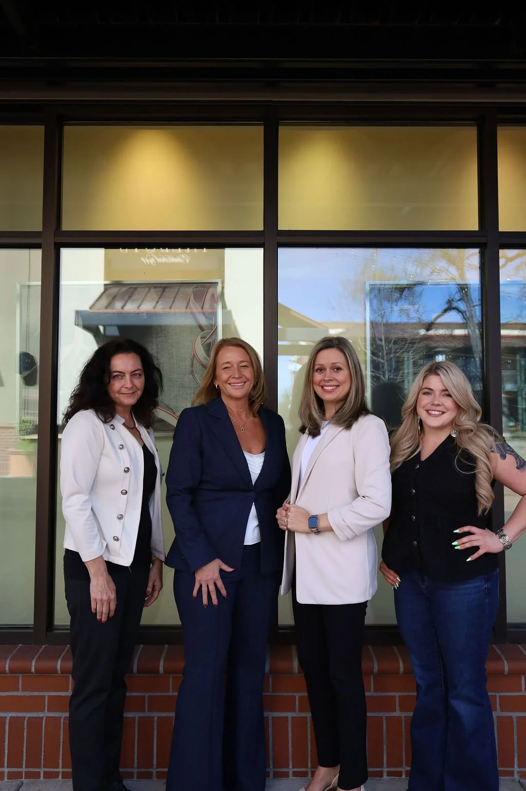 Four women in business attire standing outside in front of a building with large glass windows, smiling for the photo.