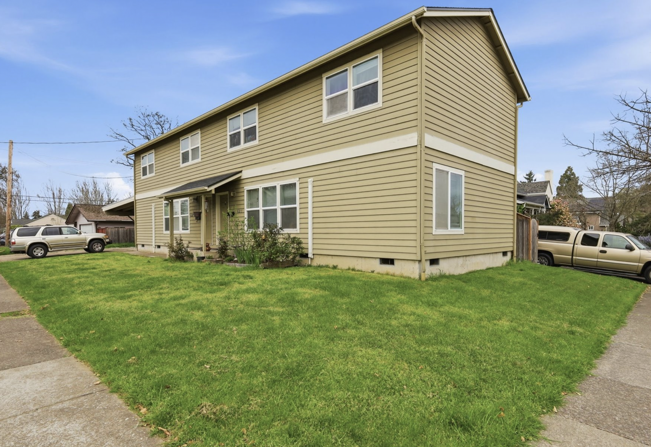 A two-story beige house with white trim and a small front porch, surrounded by a green lawn with sidewalk paths, in a suburban neighborhood with parked cars and trees.