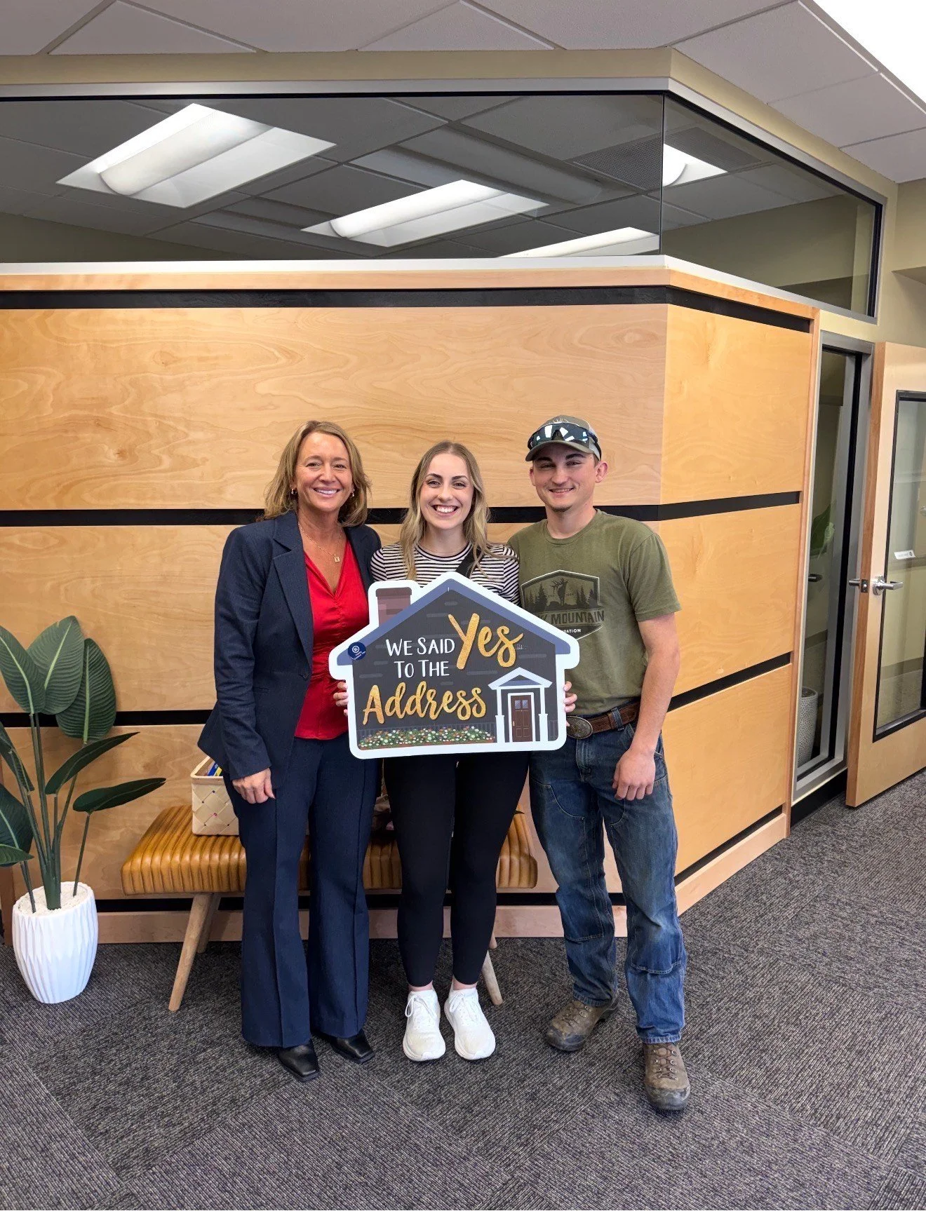Three people standing inside an office, holding a sign that reads "We said Yes to the Address," smiling and celebrating a home buy or sale. The background features a wooden partition, an indoor plant, and office furniture.