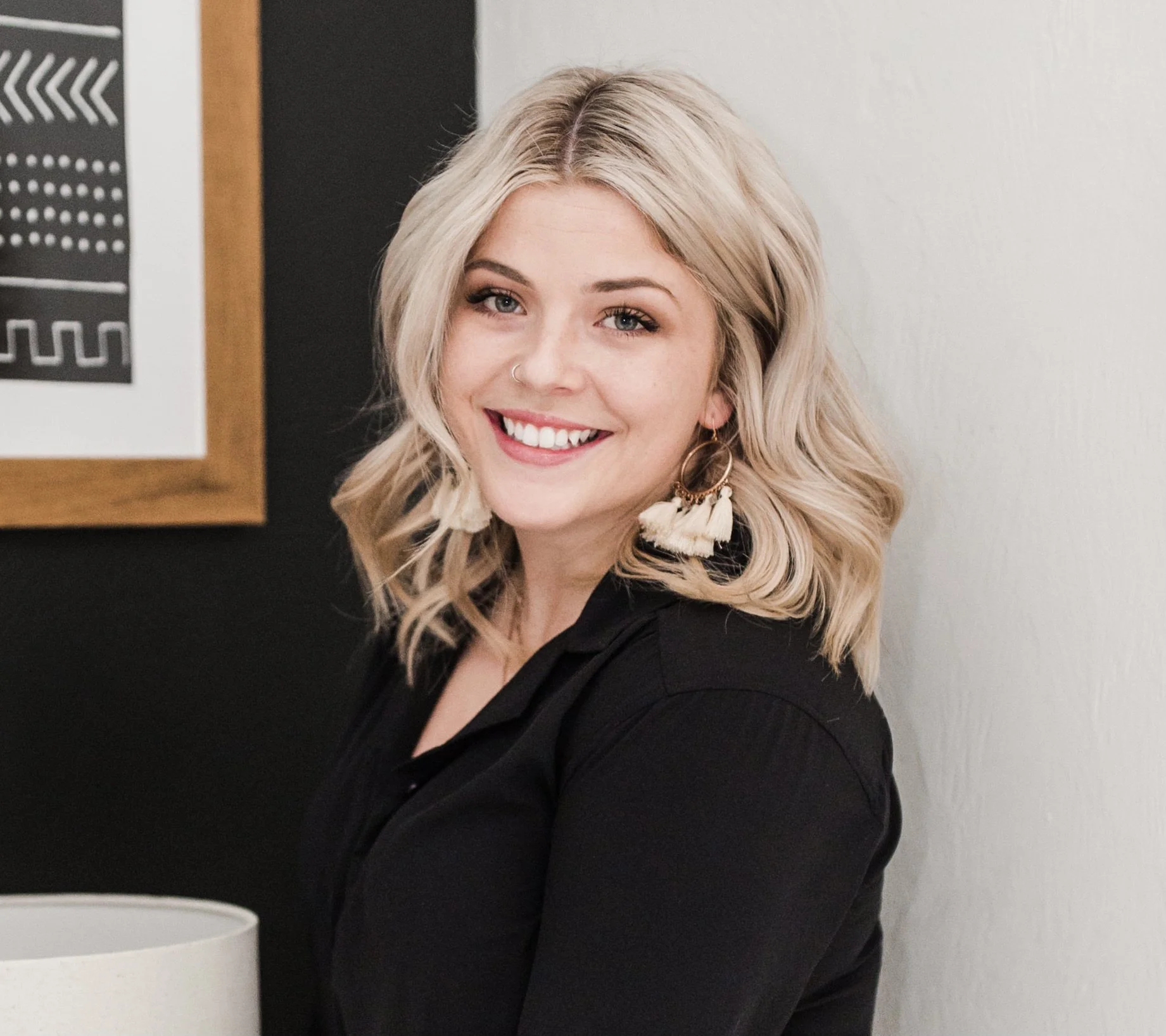 A smiling woman with blonde, wavy hair and large earrings standing indoors with a black and white wall art behind her.