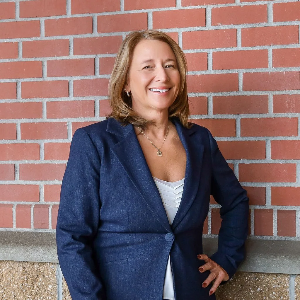 A woman with shoulder-length light brown hair smiling, dressed in a dark blue blazer and white blouse, standing against a red brick wall.