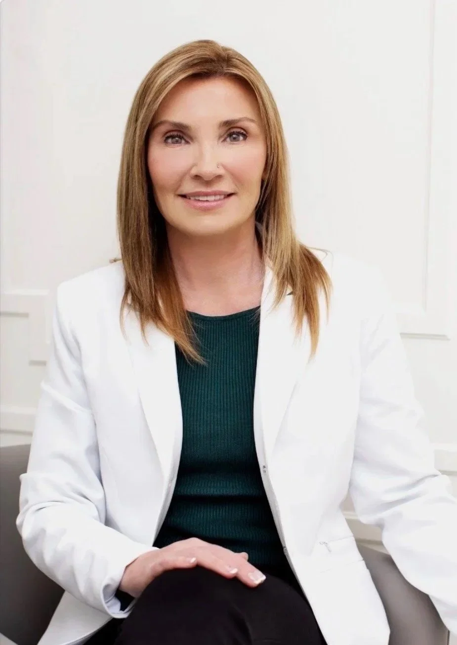 A woman with shoulder-length light brown hair wearing a white blazer and dark green top, sitting indoors against a neutral background.