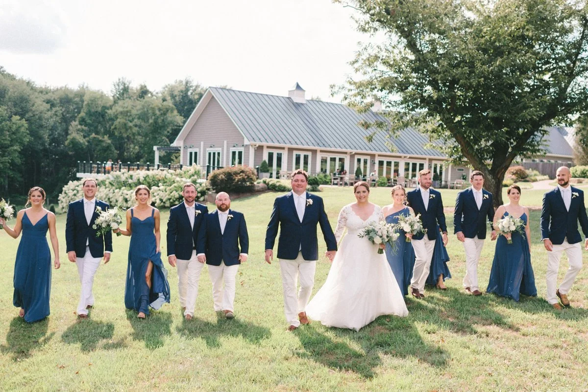 A wedding party walking outdoors on a grassy lawn, with a large house and trees in the background. The group includes bridesmaids in blue dresses, groomsmen in navy jackets and white pants, and a bride in a white wedding gown holding a bouquet, along with a groom.