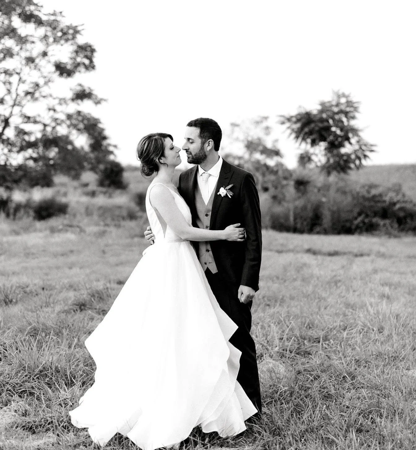 A black-and-white photograph of a bride and groom standing in a grassy field, gazing into each other's eyes, with trees in the background.