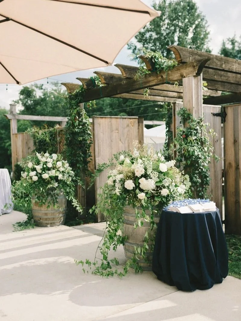 Decorative outdoor setting with large floral arrangements in wooden barrels, green vines, wooden structure, beige umbrella, and a small table with guest book menus.