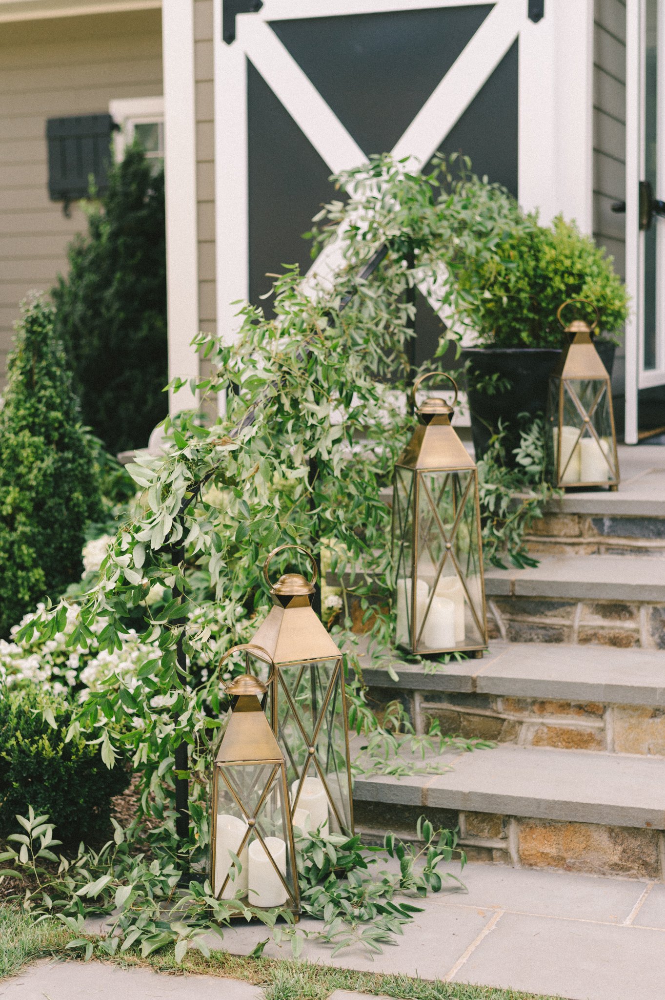 Decorative outdoor porch with greenery, lanterns, and candles, featuring stone stairs and a barn door with a geometric pattern.