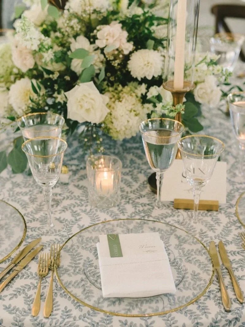 Elegant wedding table setting with gold cutlery, glassware, white and green floral centerpiece, candle, and table number card.