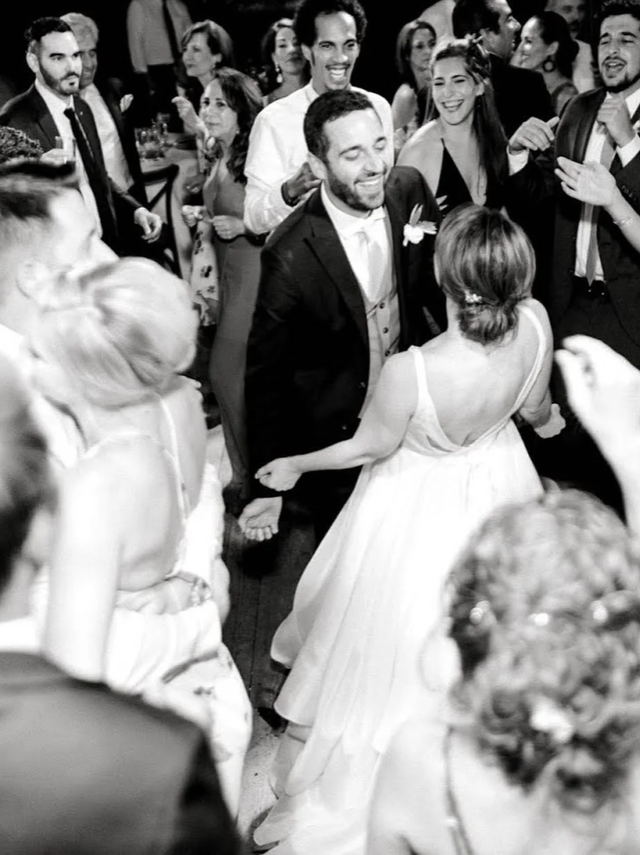 A black and white photo of a wedding reception dance, with a bride and groom holding hands and dancing, surrounded by smiling guests wearing formal attire.
