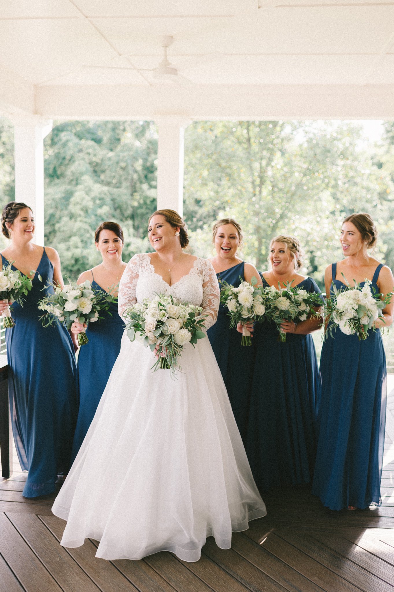 A bride in a wedding dress holding a bouquet surrounded by five bridesmaids in navy blue dresses holding bouquets standing on a wooden floor with a green outdoor background.