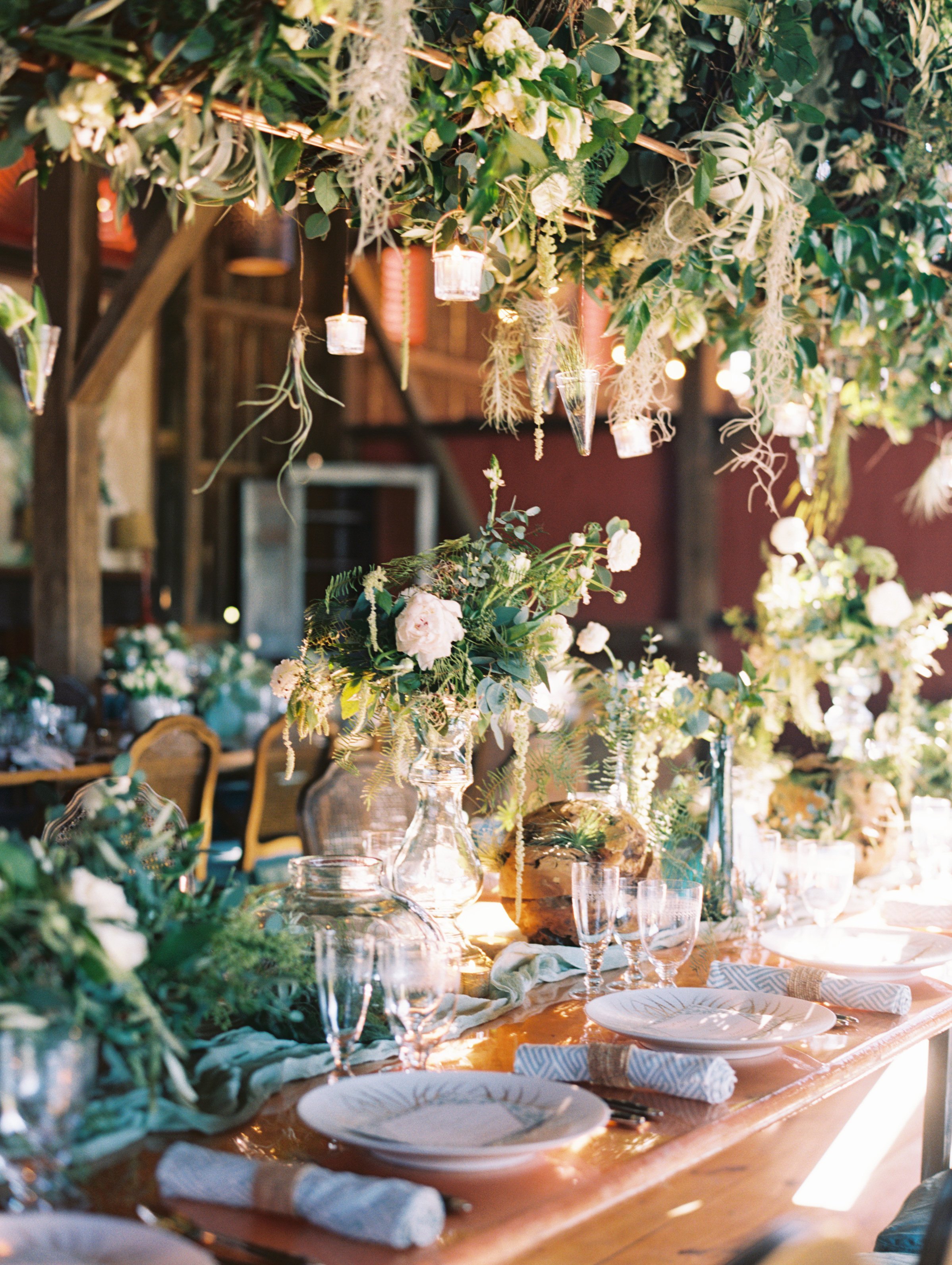 A decorated event table with floral arrangements, greenery, candles, glassware, plates, and napkins in a rustic setting.