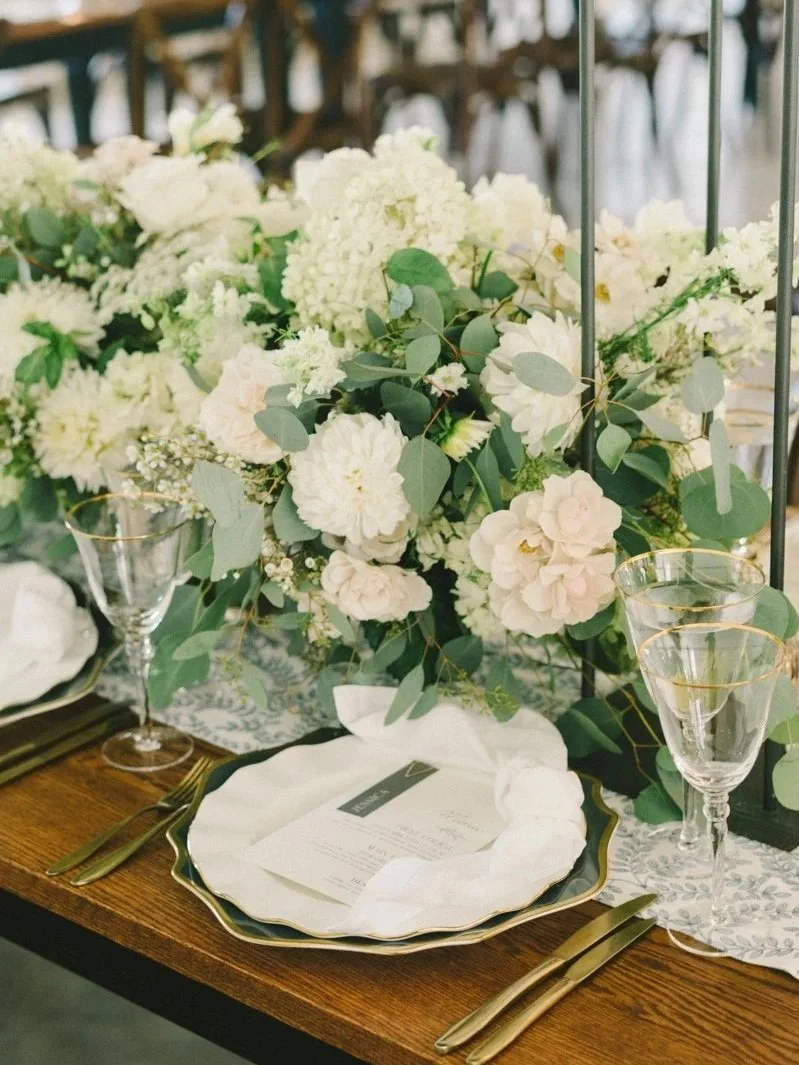 Elegant wedding table setting with a floral centerpiece, gold-rimmed glassware, and a white plate with a menu, on a wooden table with a lace table runner.