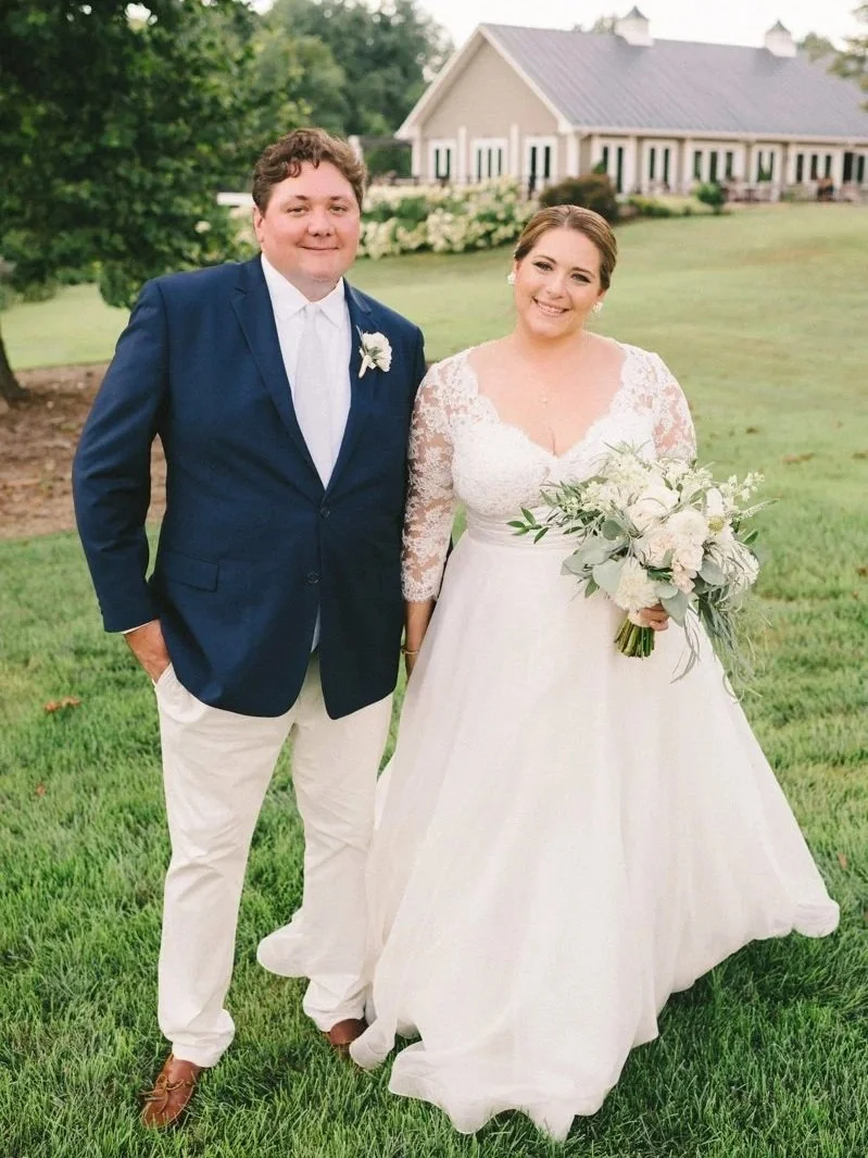 A smiling bride in a lace wedding dress holding a bouquet of white flowers, standing next to a groom in a navy blazer, white shirt, and light-colored pants, outdoors on a grassy lawn with a white house and green trees in the background.