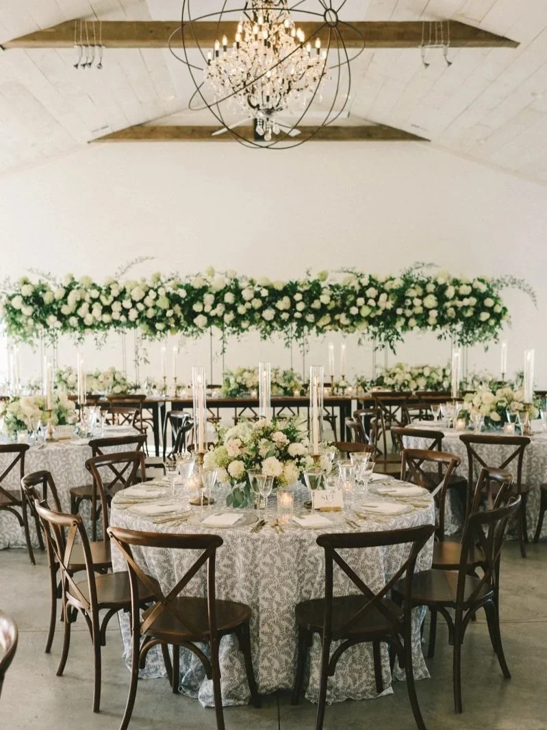 Elegant wedding or event reception with round tables, floral centerpieces, candles, and white floral decor on a long hanging arrangement, in a venue with a chandelier and a high arched ceiling.
