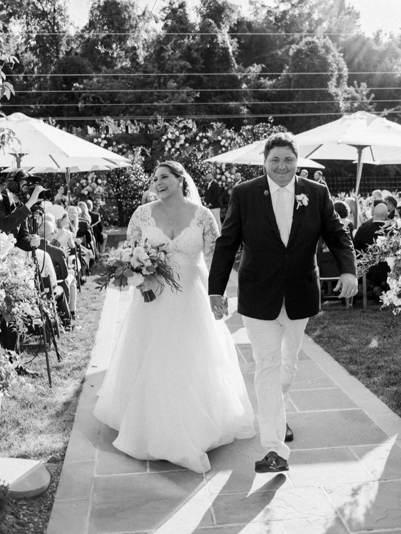A bride and groom walking hand in hand down an outdoor aisle during their wedding ceremony. The bride is wearing a white wedding gown and holding a bouquet, smiling and laughing. The groom is dressed in a black jacket, white shirt, and white pants, also smiling. Guests are seated on either side under umbrellas, with flowers and trees in the background.