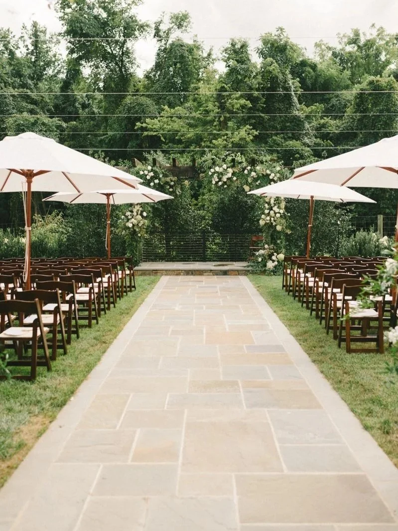 Outdoor wedding setup with a stone pathway, wooden chairs on both sides, large white umbrellas, and a floral arch at the end, surrounded by green trees.