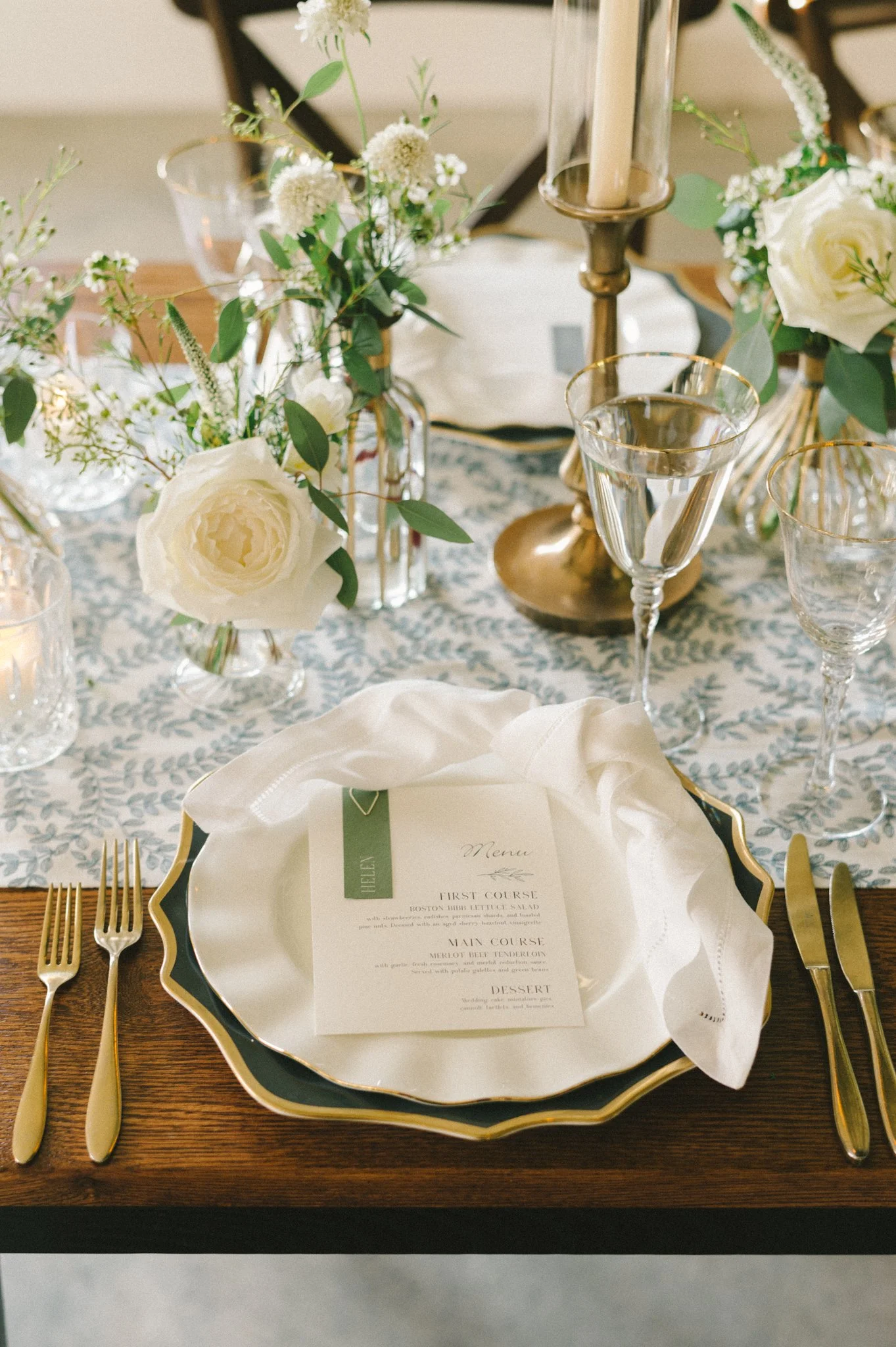 Elegant table setting with a decorative plate, gold utensils, a menu card, floral arrangement with white roses and greenery, glassware, and a gold candlestick.