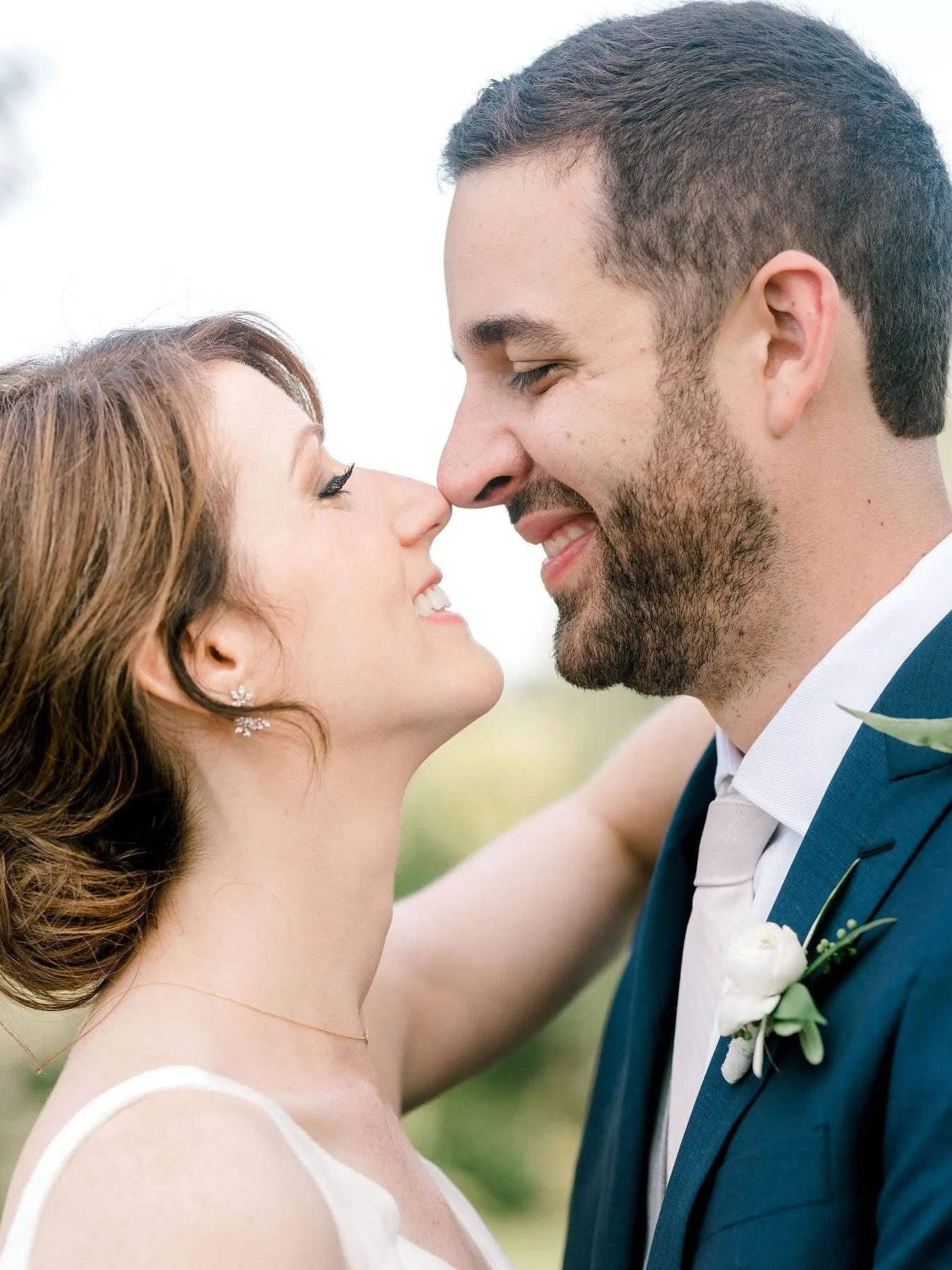A bride and groom are touching their noses together, smiling, outdoors during daytime, with the bride in a white wedding dress and the groom in a blue suit with a white shirt and boutonniere.