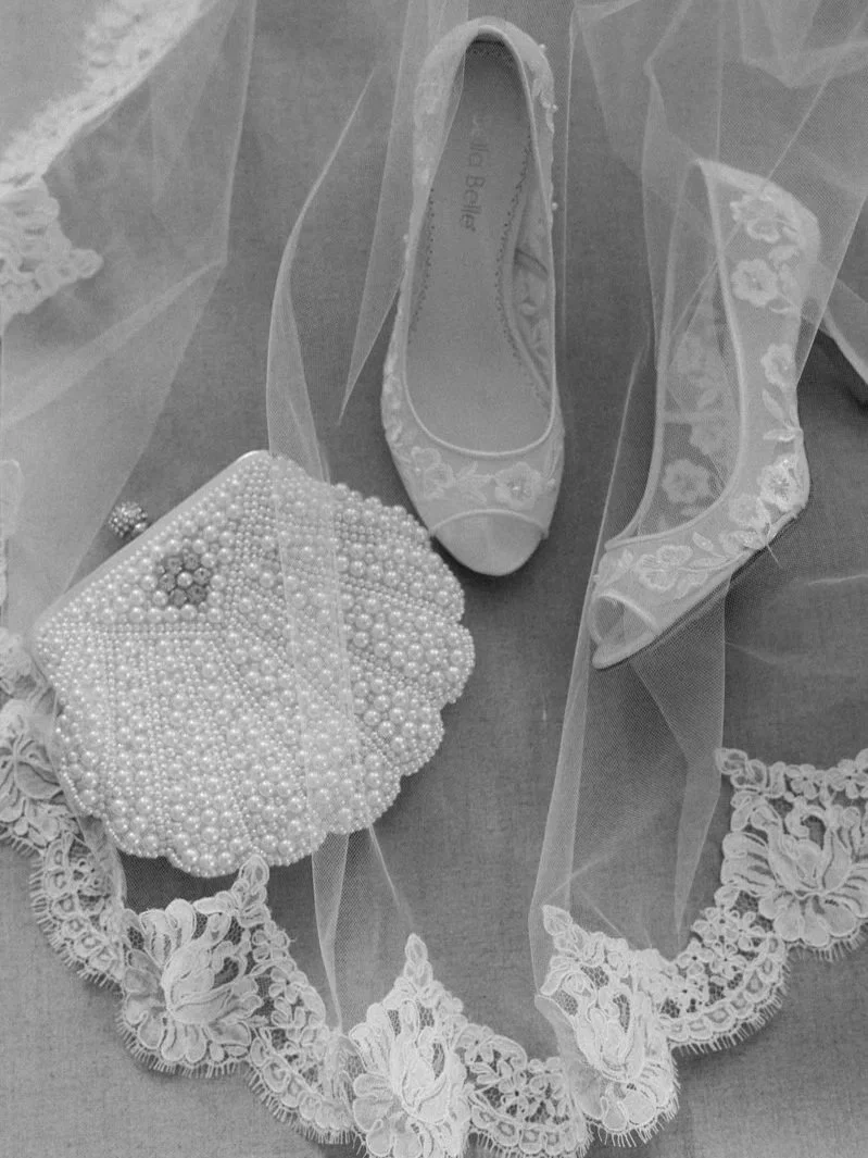 A wedding veil with floral embroidery, a single open-toe satin shoe with embroidery on the toe, and a pearl-embellished clutch purse.