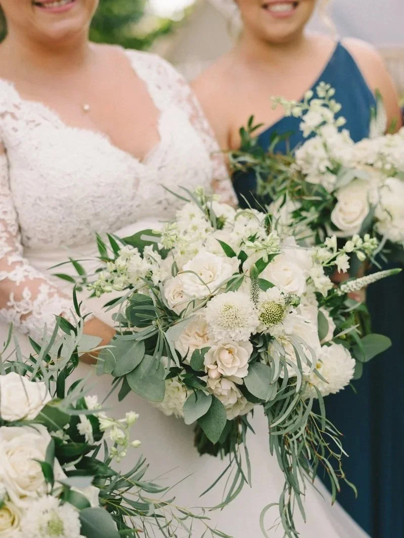 Two women holding white floral bouquets, one wearing a lace wedding dress, and both smiling.