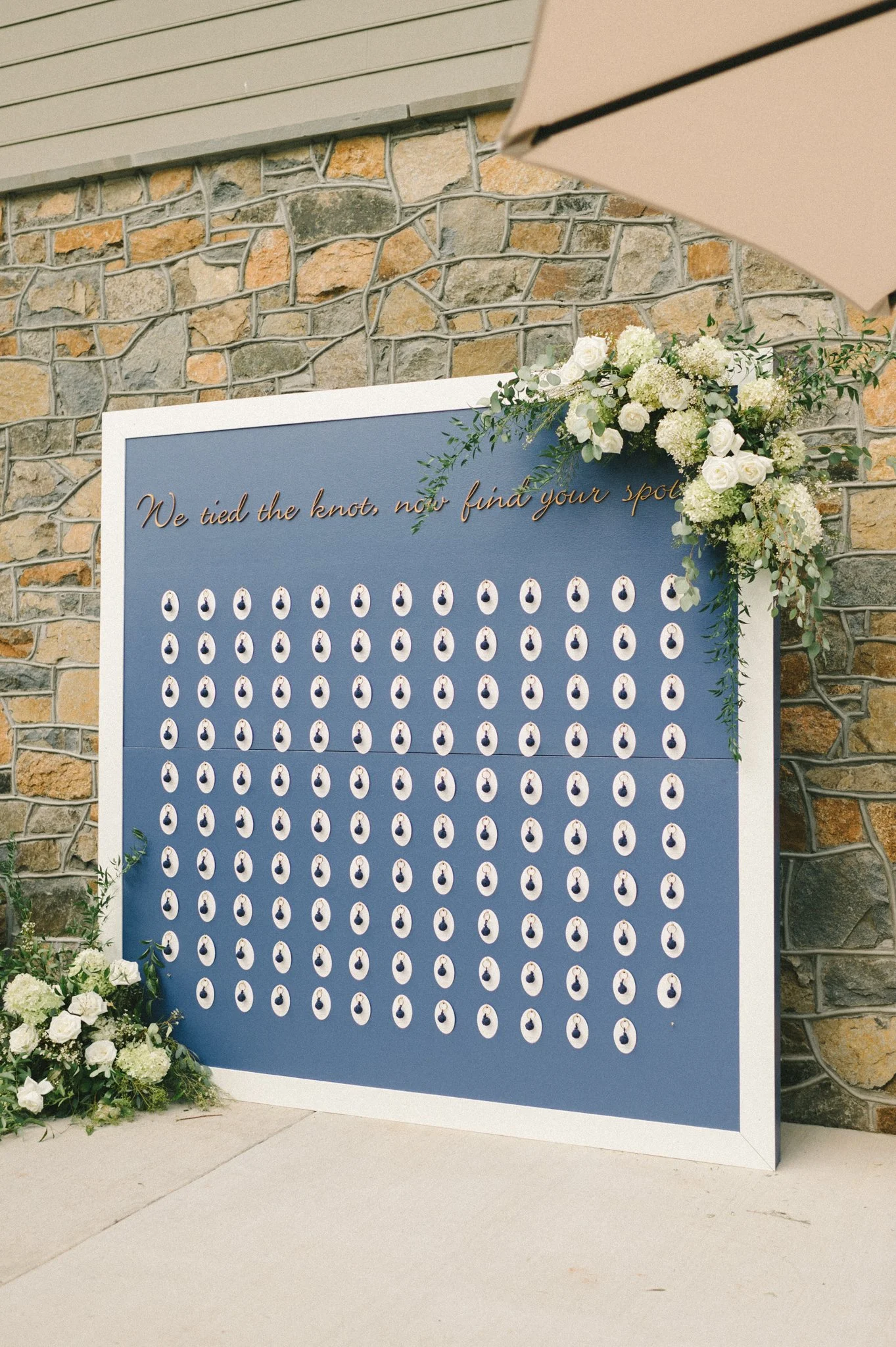 A large display board decorated with white flowers and greenery, featuring a quote and rows of small ceramic or porcelain hanging objects, set against a stone wall and partially covered by a building or awning.