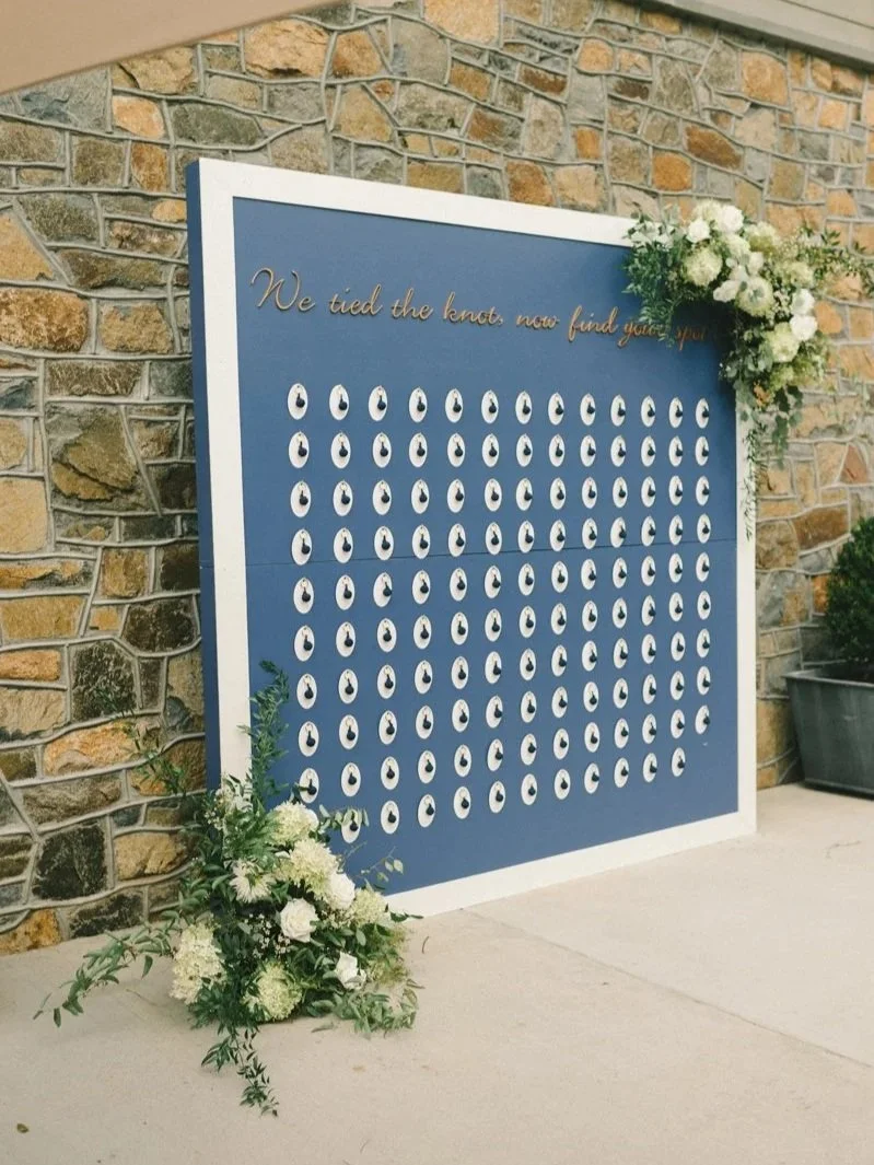 A wedding seating chart with white circular tags arranged on a blue background. There are floral arrangements at the top right and bottom left corners. A sign with the text "We tied the knot, now find your spot" is at the top.