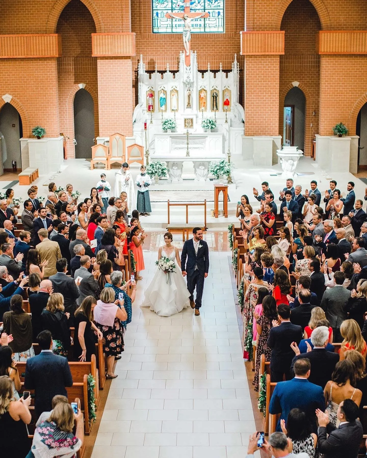 A bride and groom walking down the aisle in a church after their wedding ceremony, surrounded by seated and standing guests taking photos and clapping.