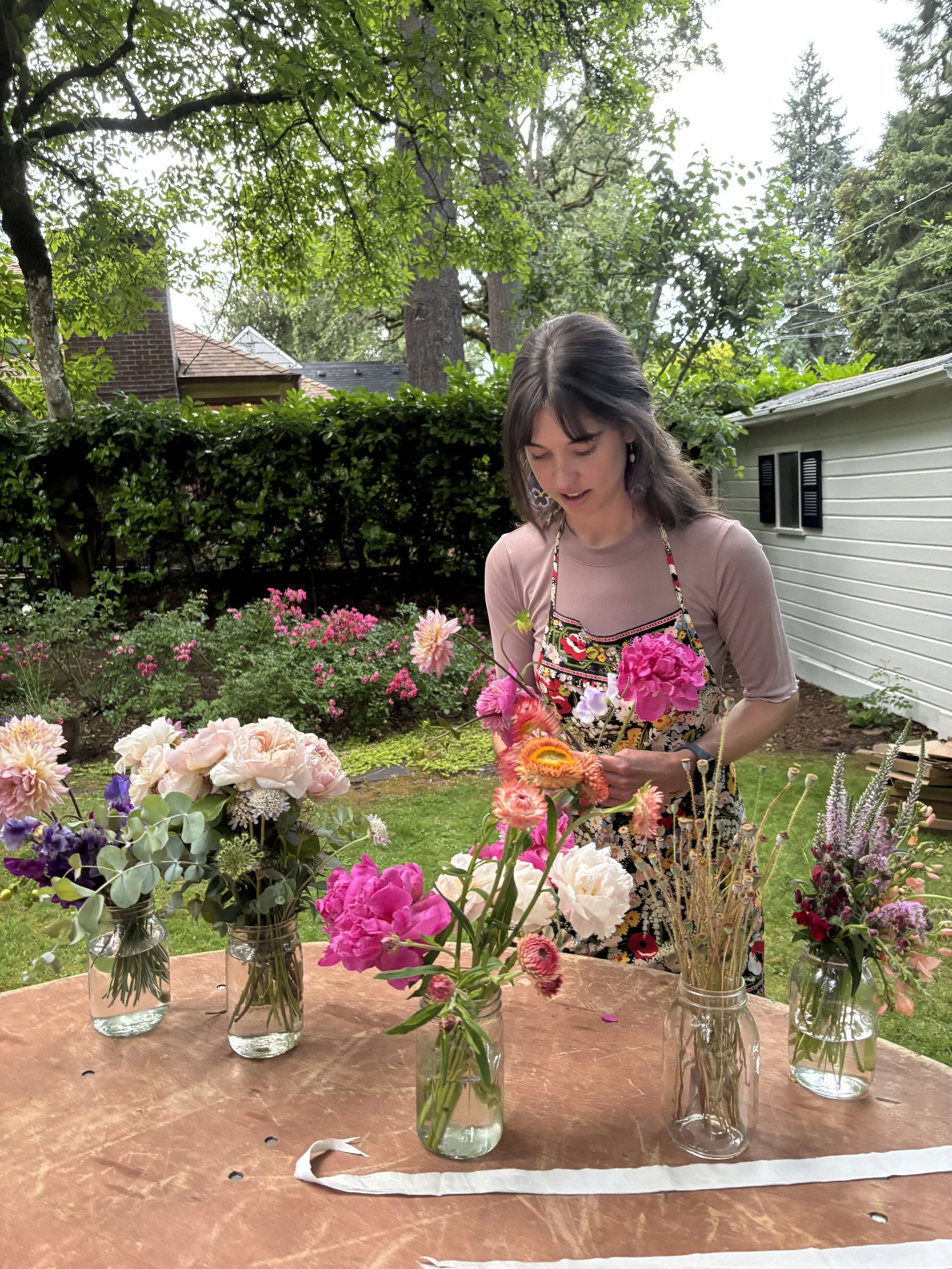 A woman arranging colorful flowers in small glass vases on a table outdoors in a garden with lush trees and a house in the background.