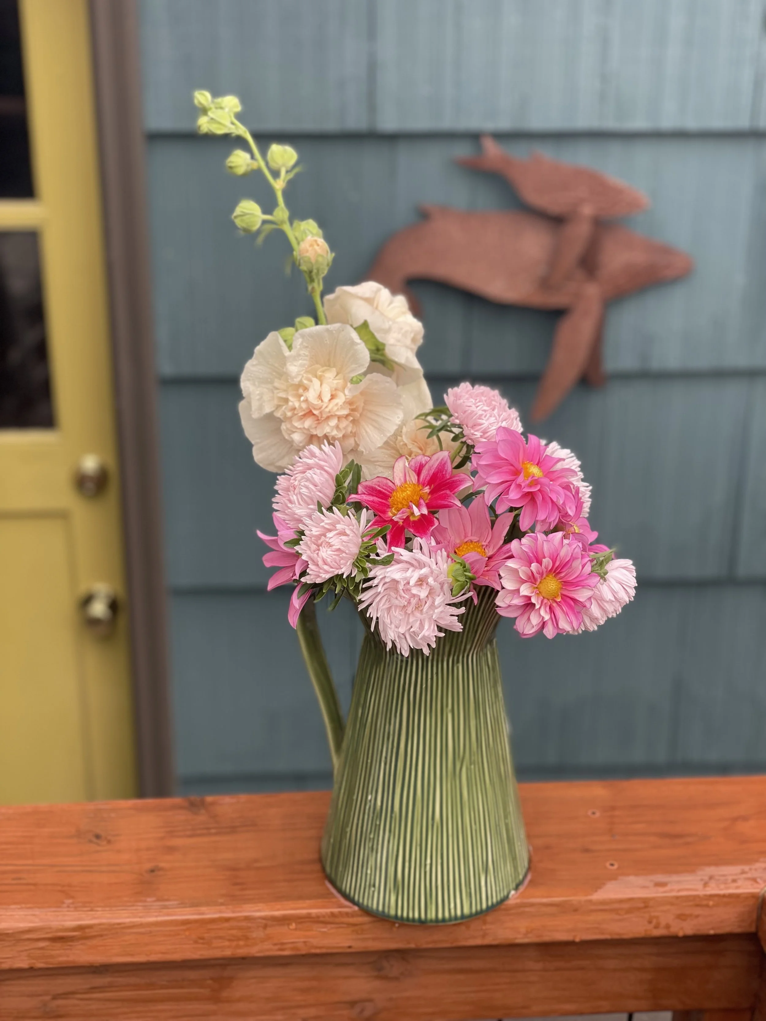 A vase with a bouquet of pink and cream flowers on a wooden table with a blue wall and wood fish wall art in the background.