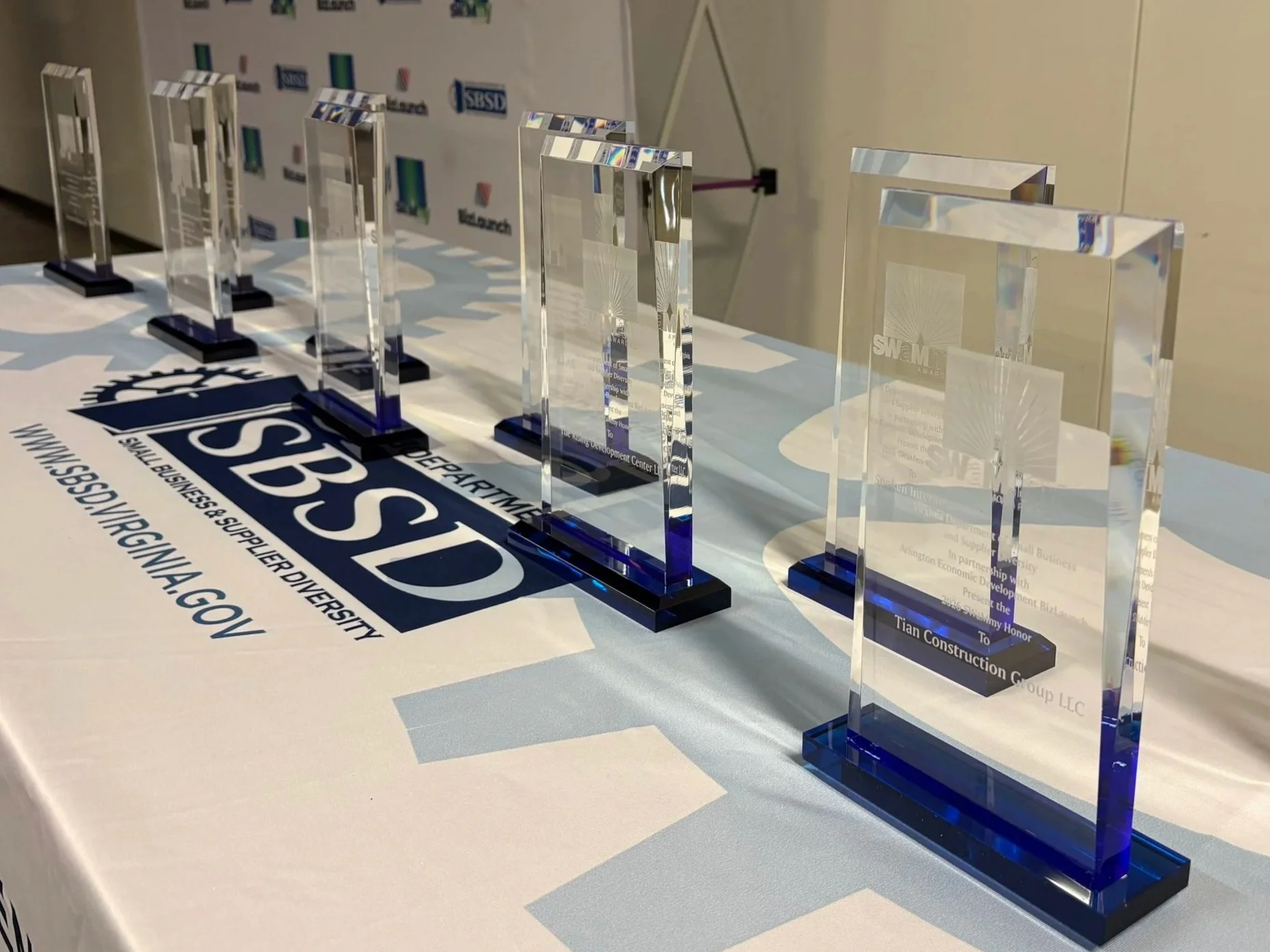 A row of crystal SWaMmy Honor awards displayed at the 2026 SWaMmy Honors ceremony in front of branded backdrops for the Virginia Department of Small Business and Supplier Diversity (SBSD) and Arlington Economic Development's BizLaunch.