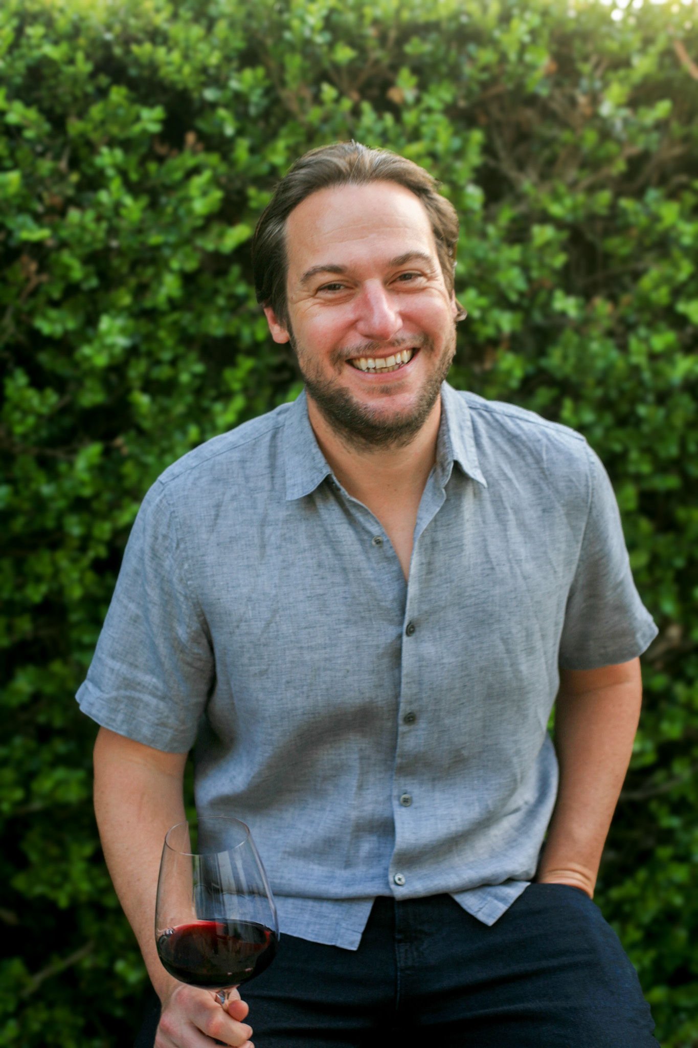 A man smiling outdoors holding a glass of red wine, wearing a light blue button-up shirt, with green bushes in the background.