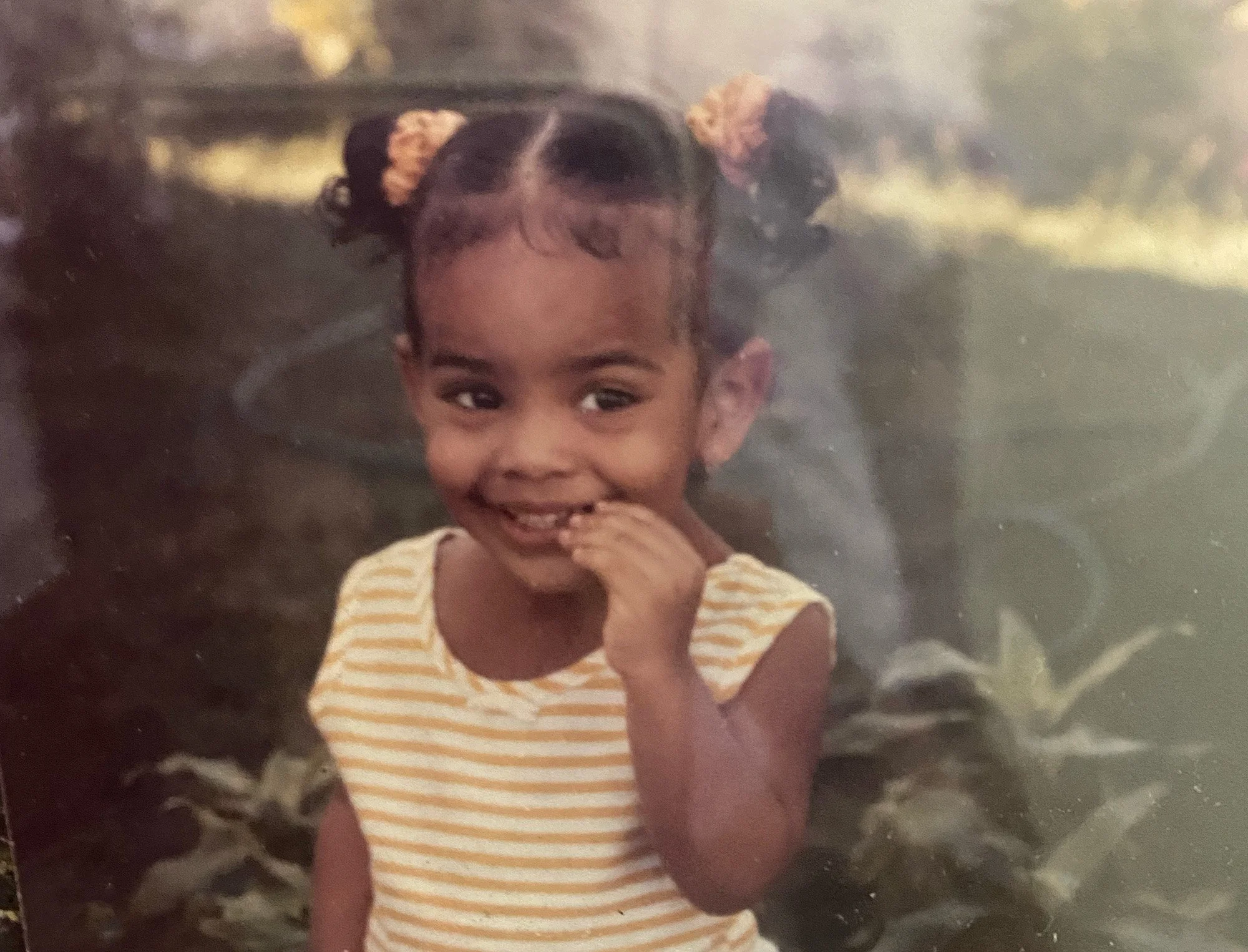 A young girl with dark hair styled in two puffy pigtails decorated with peach-colored hair ties, smiling and holding a finger to her mouth, wearing a yellow and white striped shirt.
