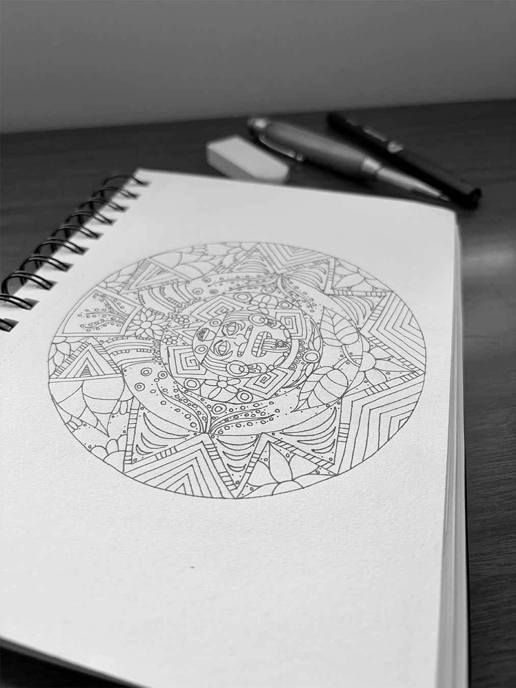 Black and white photograph of a spiral-bound sketchbook open on a desk, displaying an intricate aztec calendar drawing with various geometric patterns and floral designs. In the background, there are a pencil, pen, and an eraser on the desk.