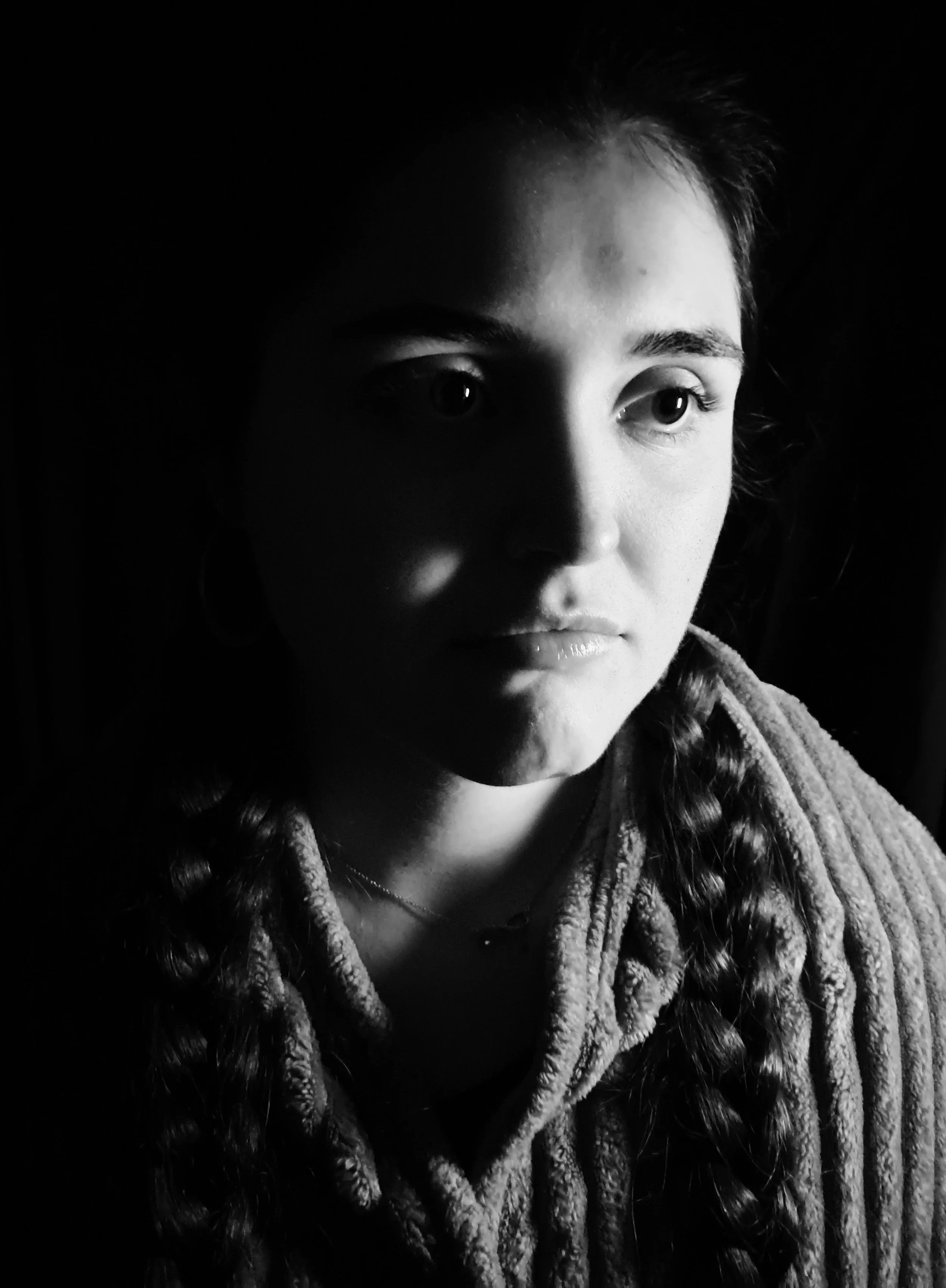 Black and white portrait of a young woman with braided hair, wearing a textured blanket, looking into the distance in a dimly lit setting.