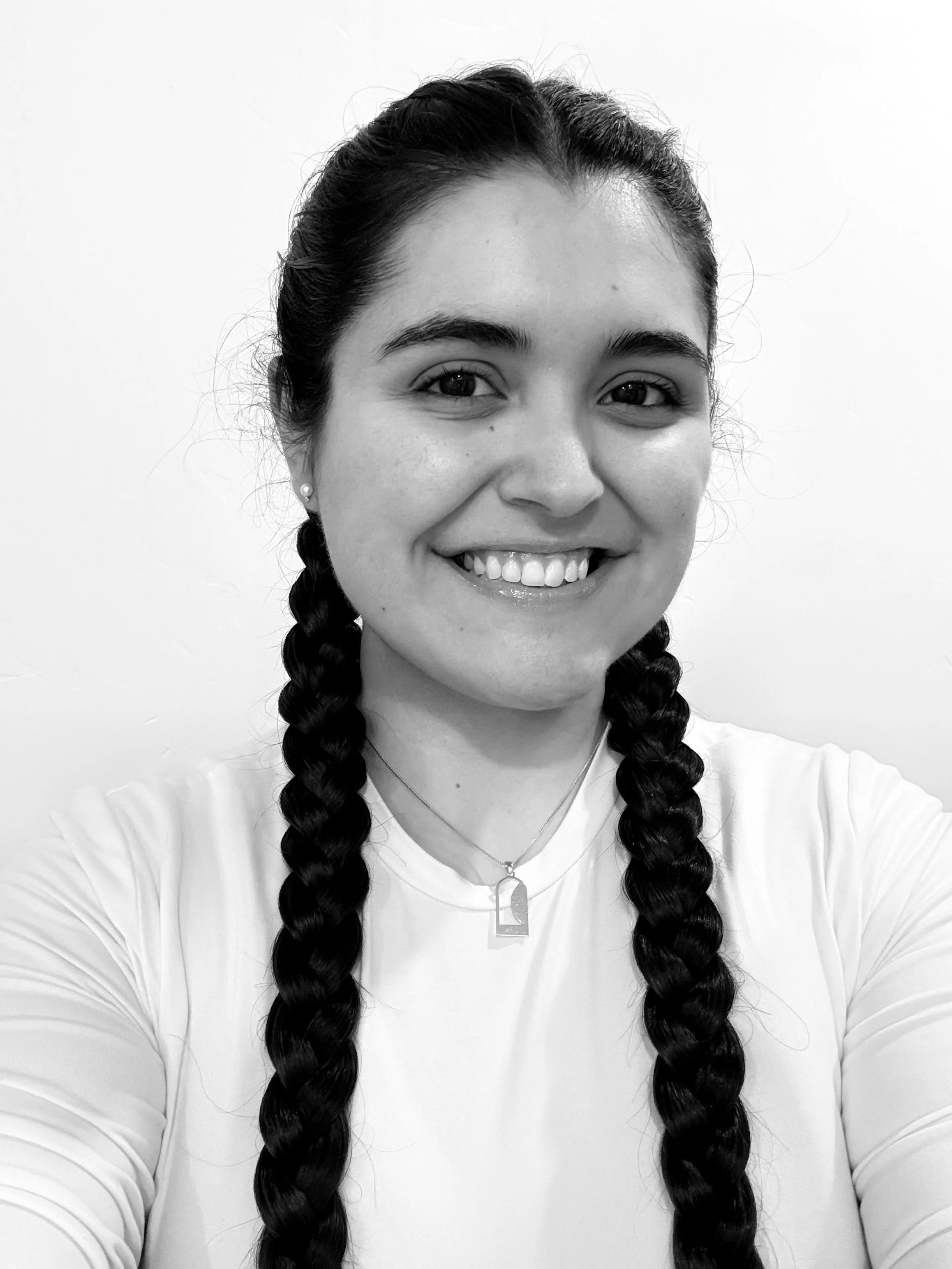 A black and white portrait of a young woman with dark hair in two braids, smiling and wearing a white shirt, small earrings, and a necklace with a rectangular pendant representing the empty tomb.