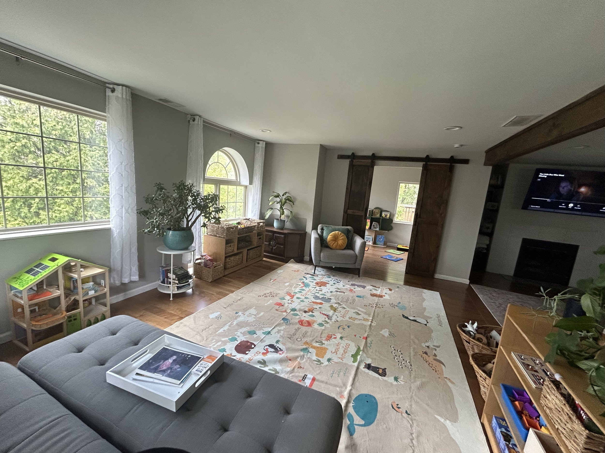 Living room with large windows, white curtains, potted plants, a children's world map rug, furniture including a gray sofa and a wooden entertainment center, and a wall-mounted TV.