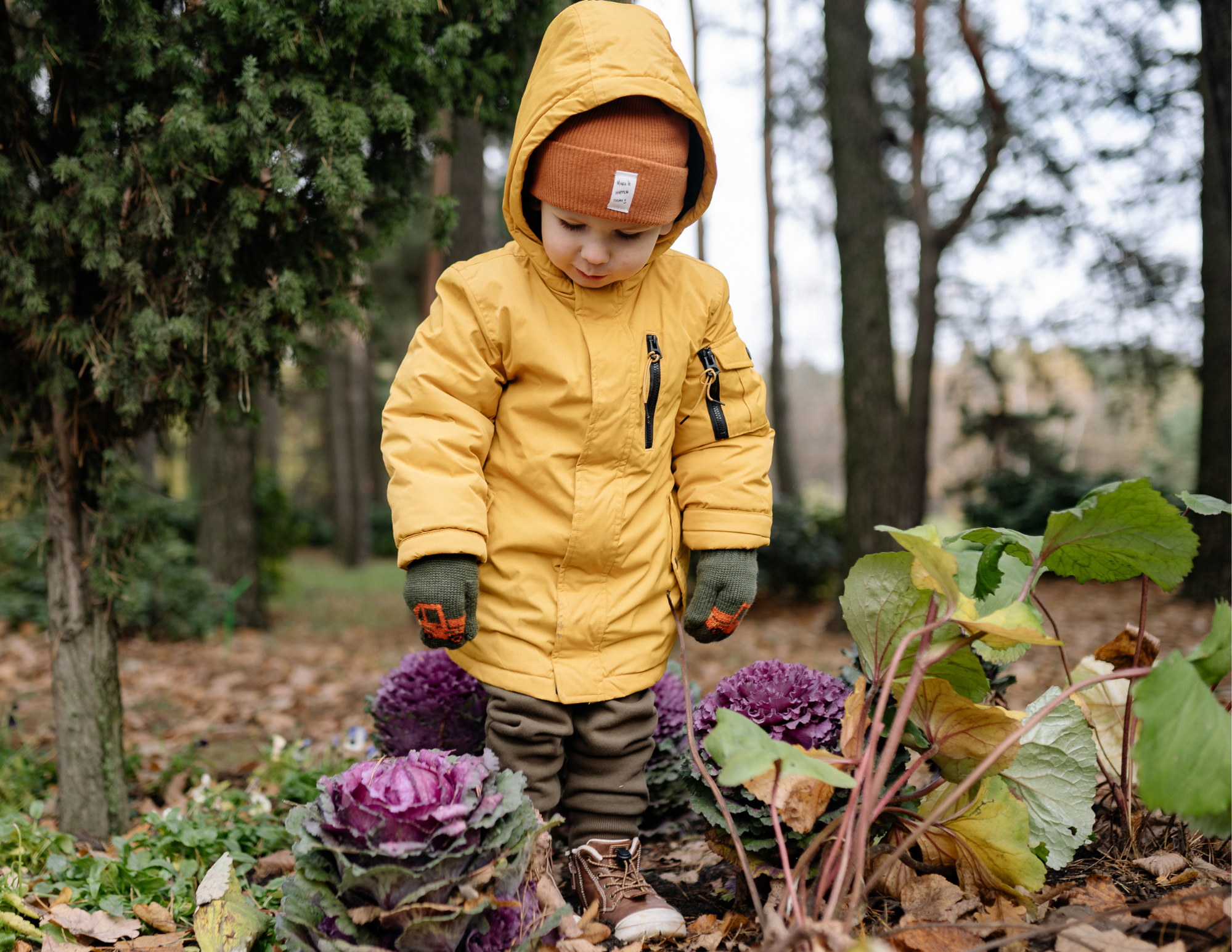 A young child wearing a yellow jacket and orange hat is in a garden surrounded by plants and flowers, with trees in the background.