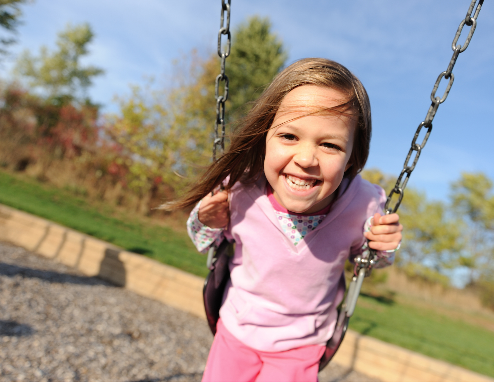 A young girl with long brown hair smiling happily on a swing at a park on a sunny day, with trees and a blue sky in the background.