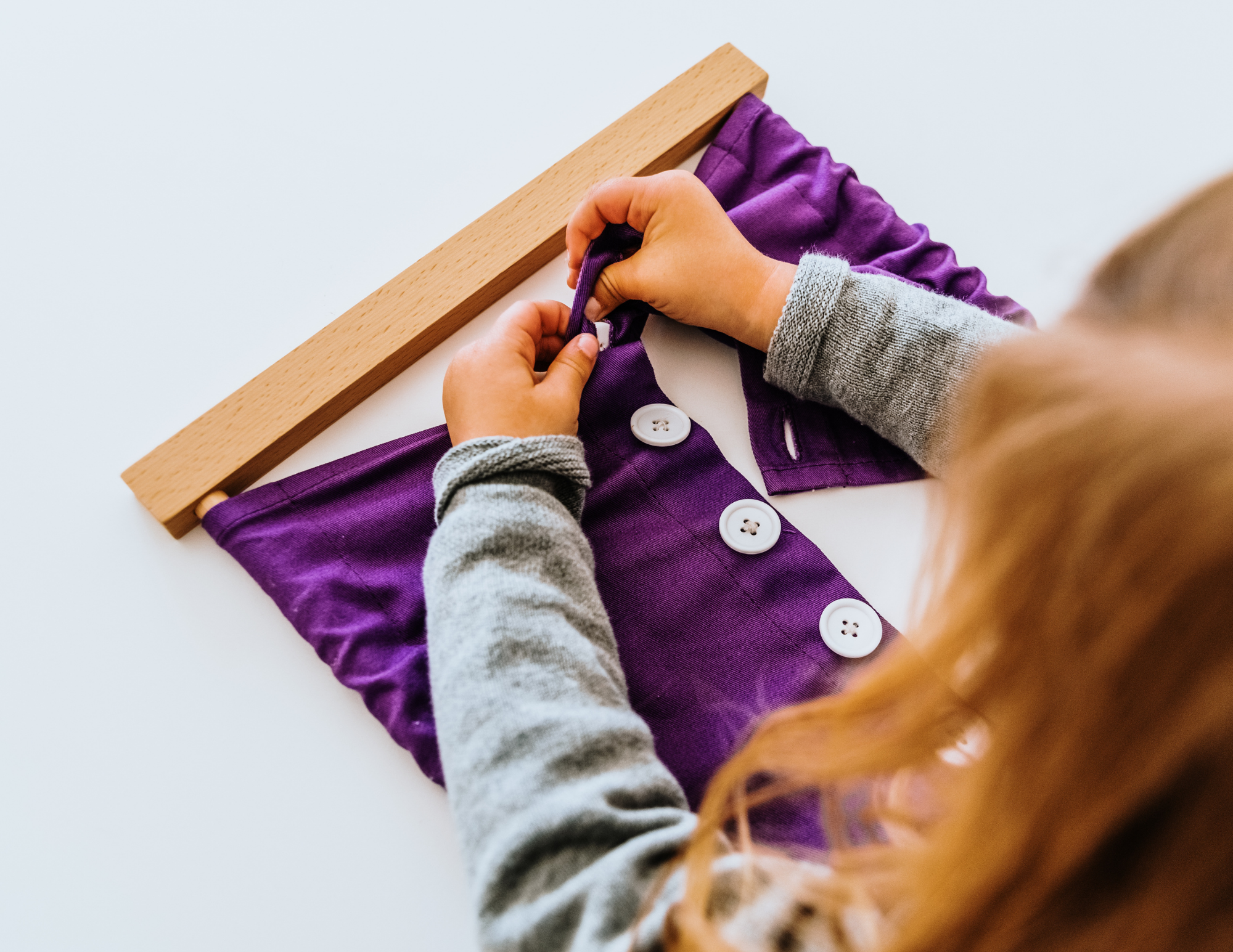 A person sewing a purple garment with white buttons, using a wooden ruler as a guide.