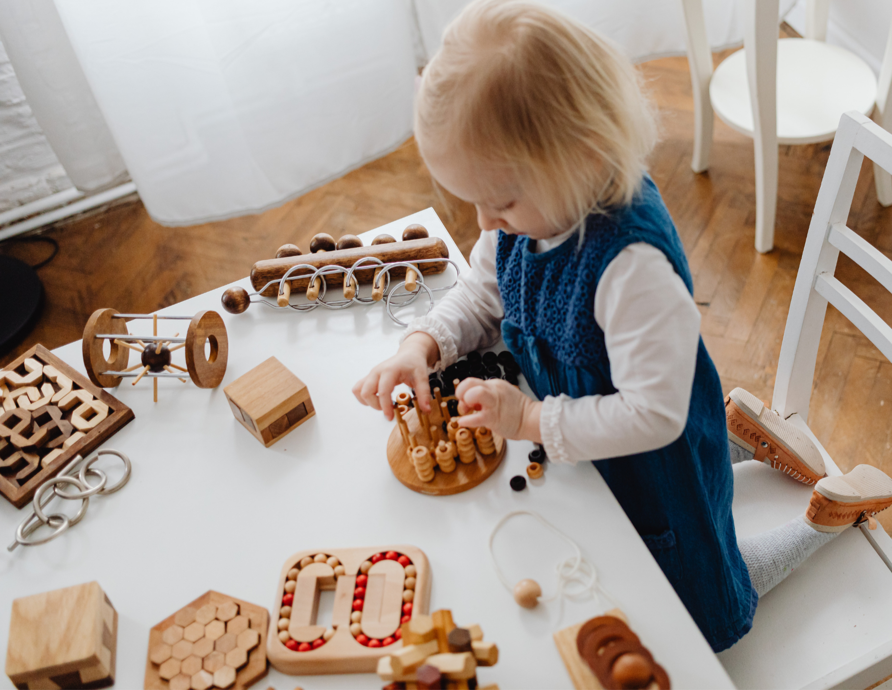 A young girl with blonde hair wearing a blue dress playing with wooden toys and building blocks on a white table in a room with wooden flooring and white chairs.