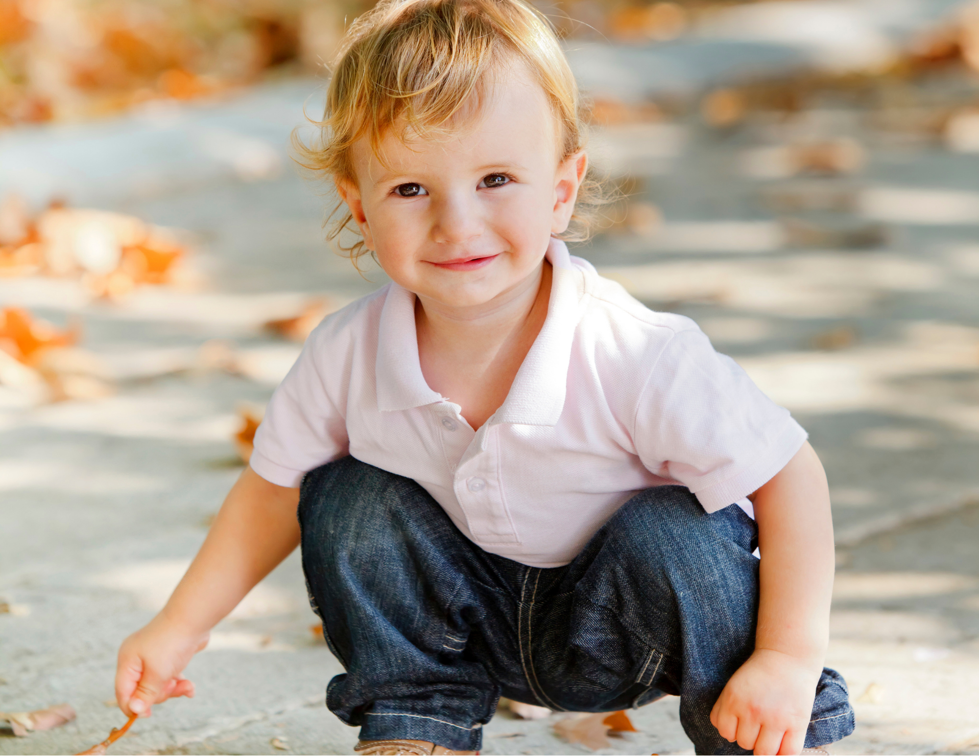 A young boy with curly blonde hair smiling and playing outdoors on a sunny day, with autumn leaves on the ground.