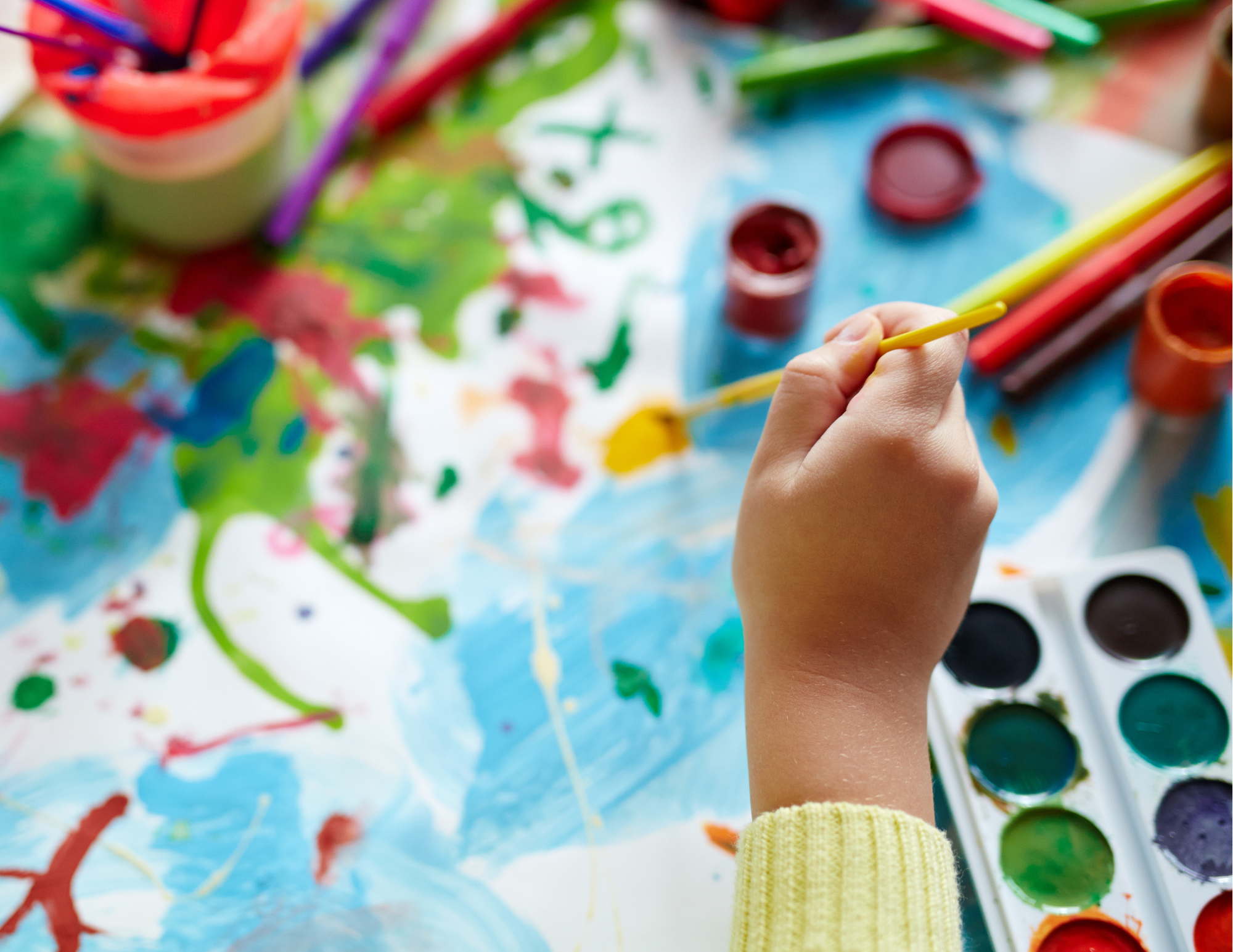 Child's hand holding a yellow paintbrush, painting with watercolors on an art table covered with colorful paint and art supplies.