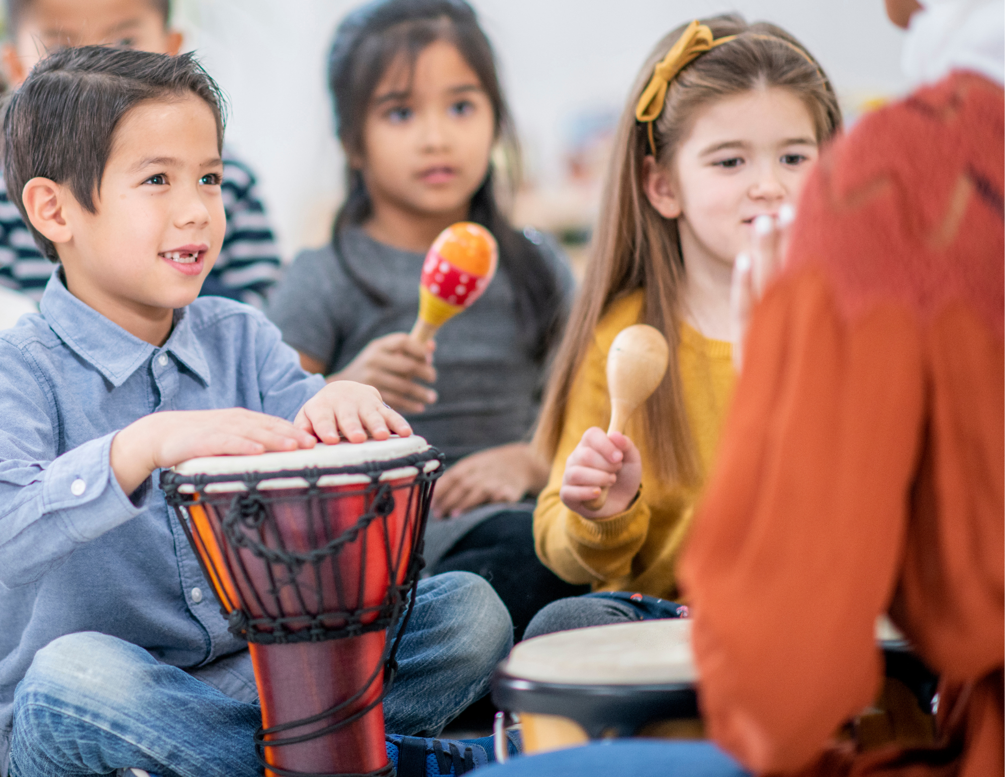 Group of children sitting and playing musical instruments, including drums and maracas, in a classroom or music room.