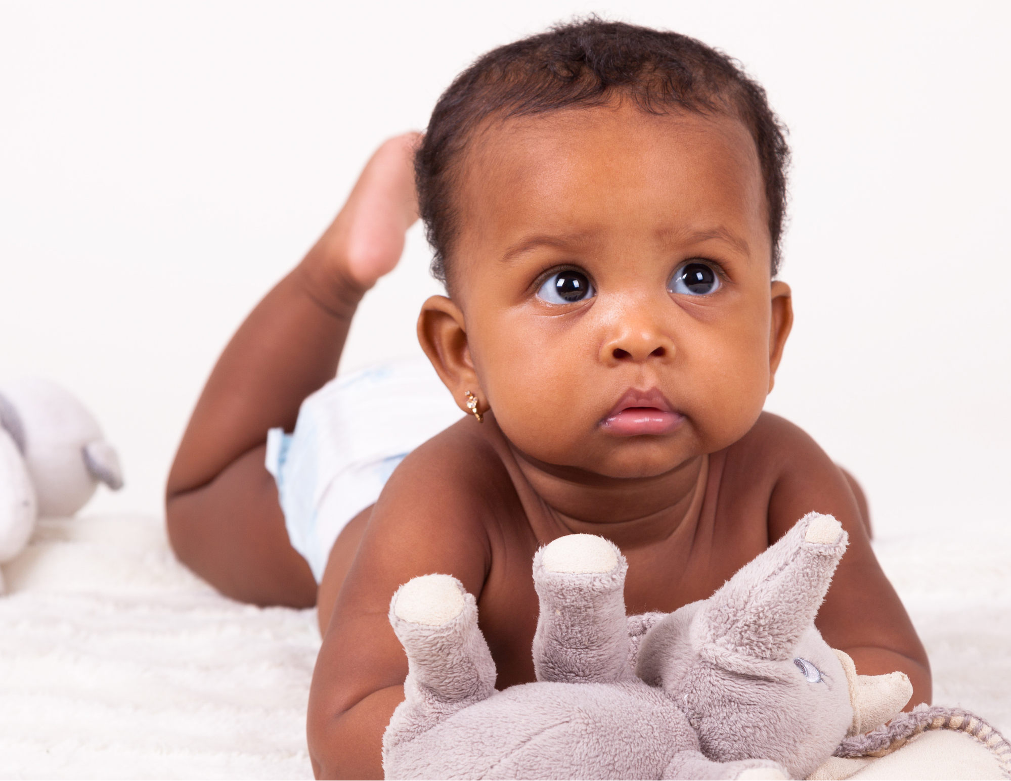 A baby crawling on a white carpet, holding a plush teddy bear, with a hand touching her head, against a plain white background.