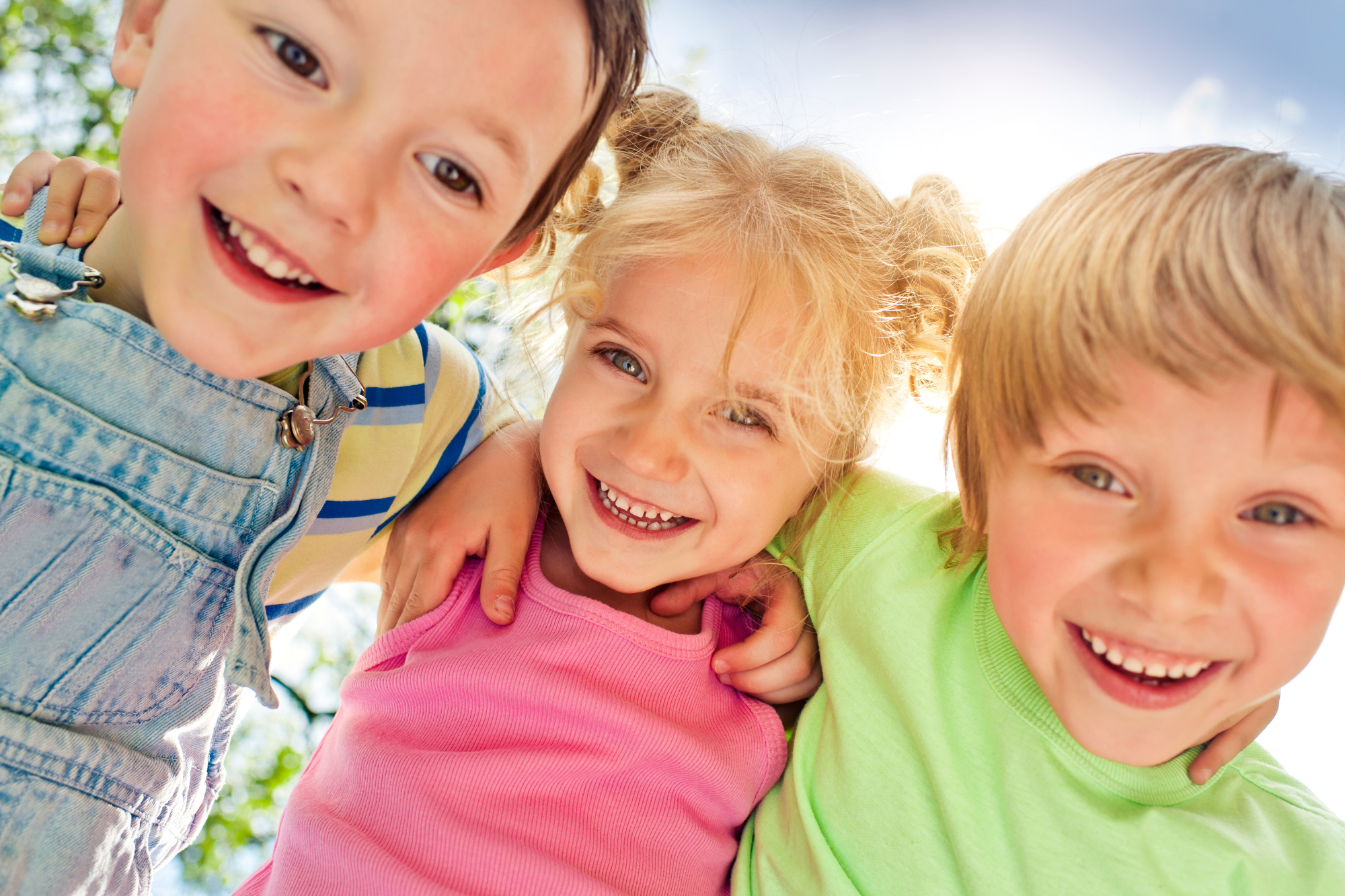 Three children smiling and looking down at the camera outdoors with trees and sky in the background.