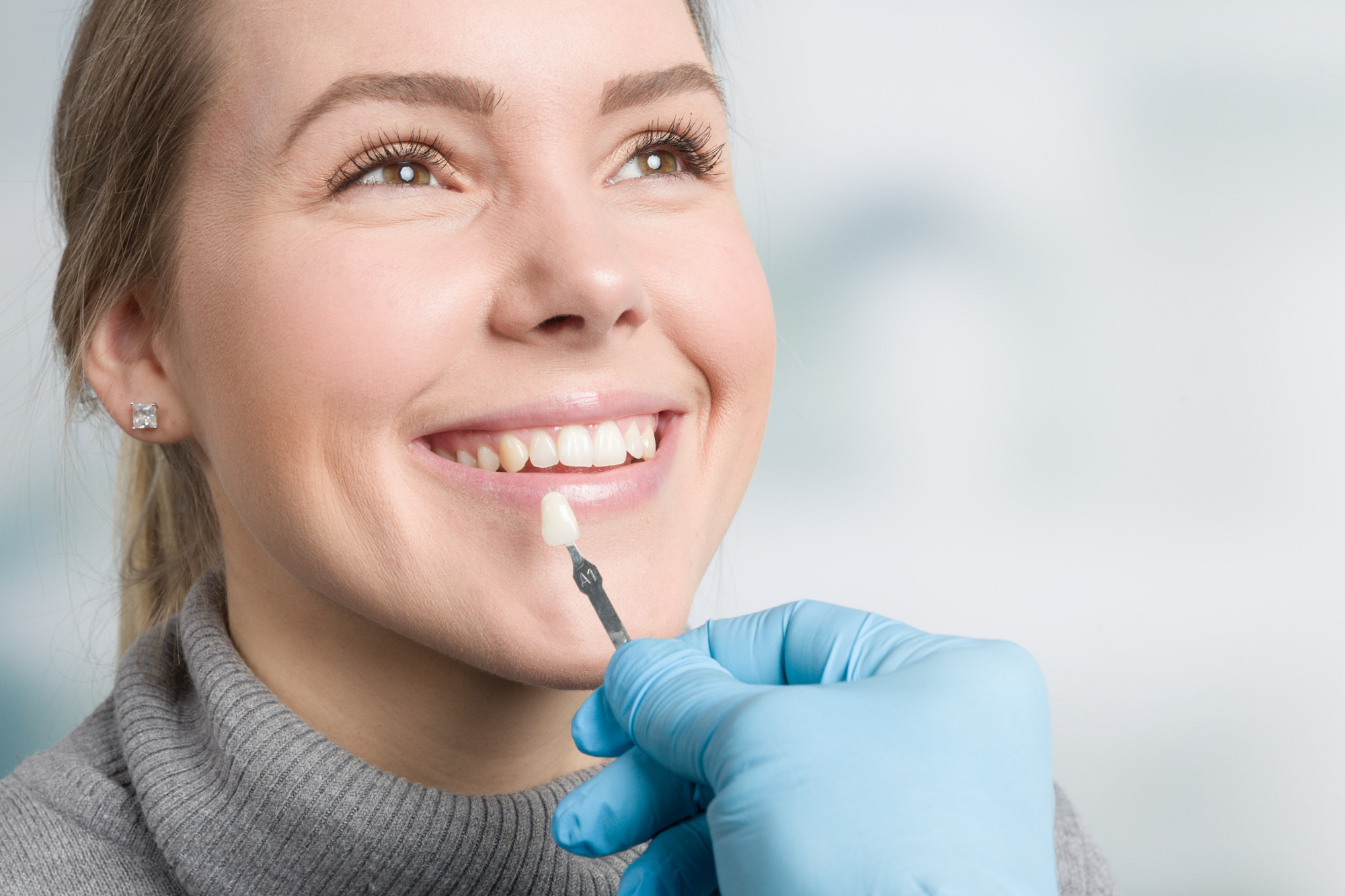 A woman at the dentist's office smiling as a dental professional in a blue glove applies a tooth-colored veneer or dental material to her upper front tooth.