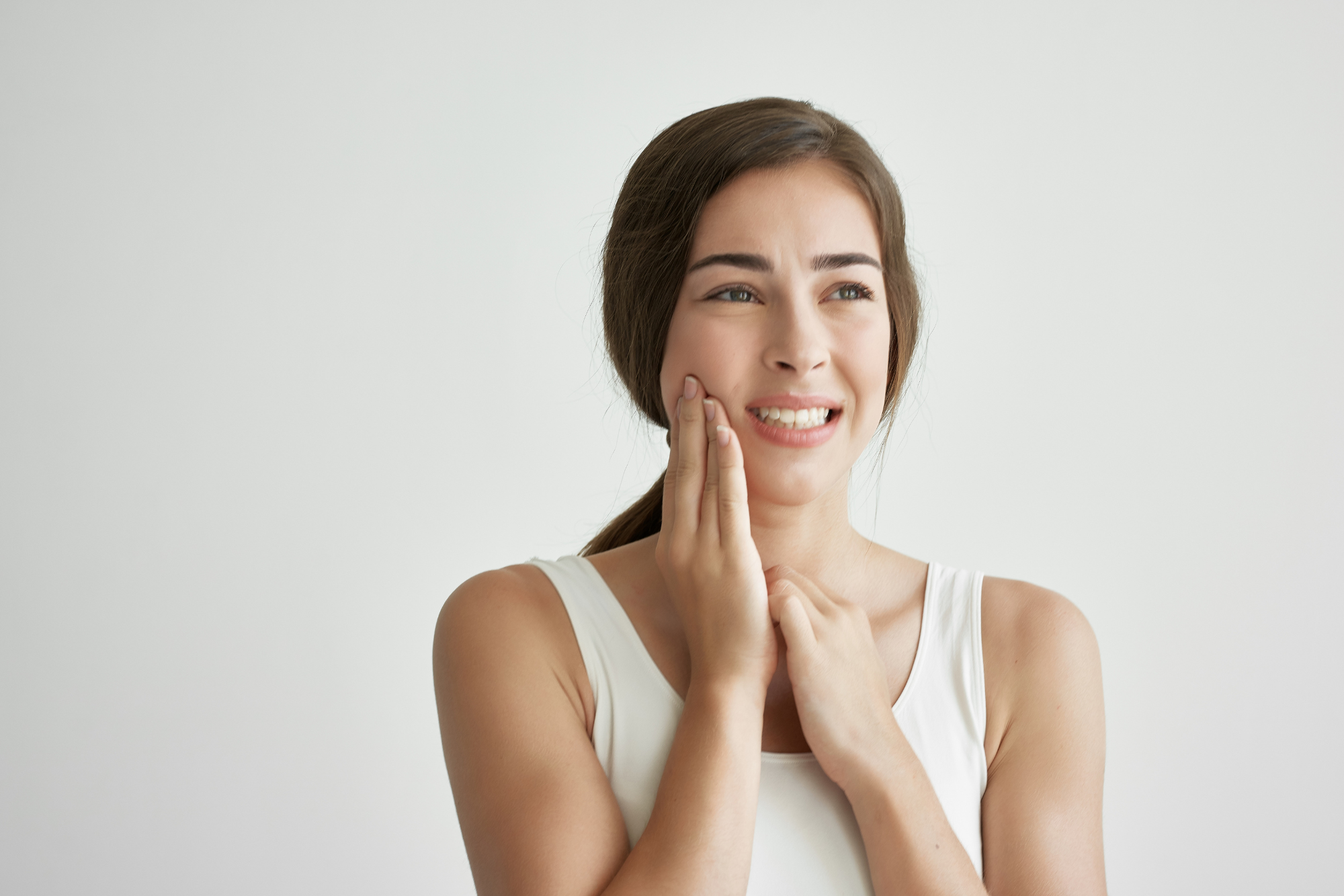 Young woman with brown hair and light skin holding her cheek and jaw, appearing to experience pain or discomfort, wearing a white tank top, against a plain white background.