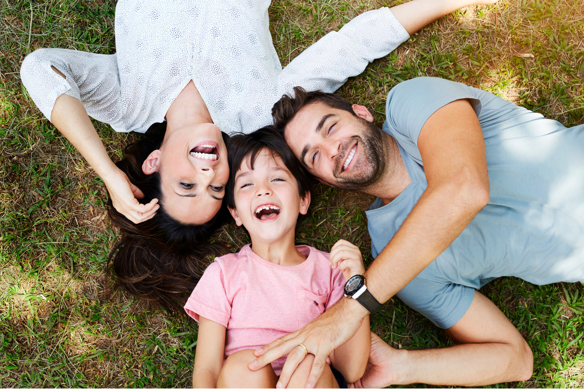 A family of three lying on grass outdoors, laughing and smiling together. The mother has dark hair, the father has dark hair and a beard, and the child has short dark hair and is wearing a pink shirt.
